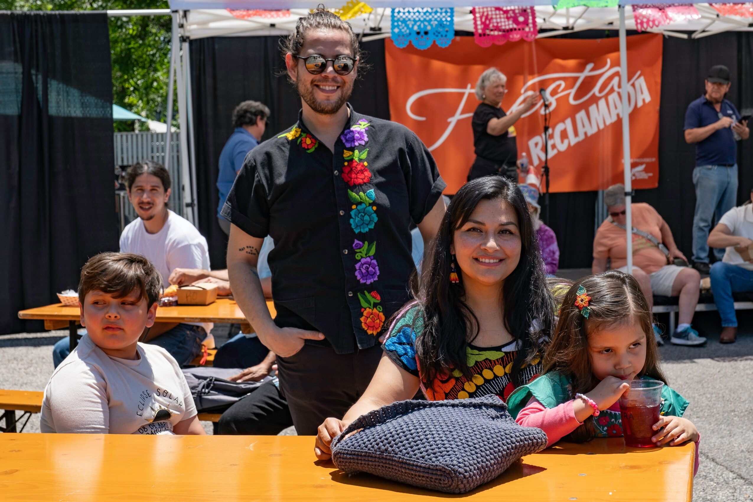 Three people sitting at a table outdoors, smiling, with a man standing behind them, in front of a booth and other people.