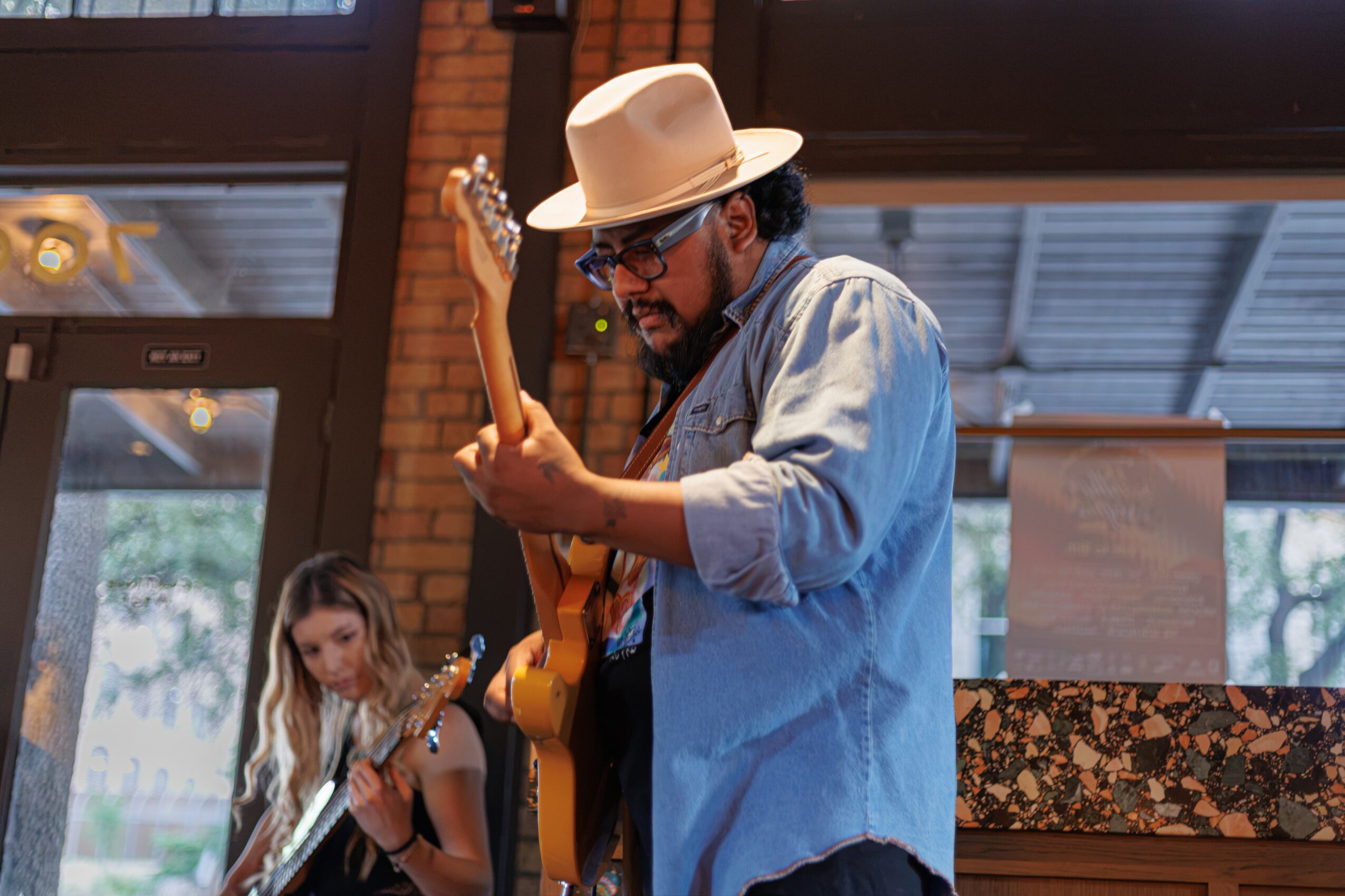 Man wearing a hat and glasses playing an electric guitar indoors with a young girl in the background.
