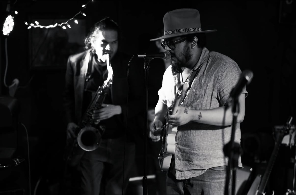 Two musicians perform on stage, one playing saxophone and the other singing and playing guitar, in a dimly lit setting.