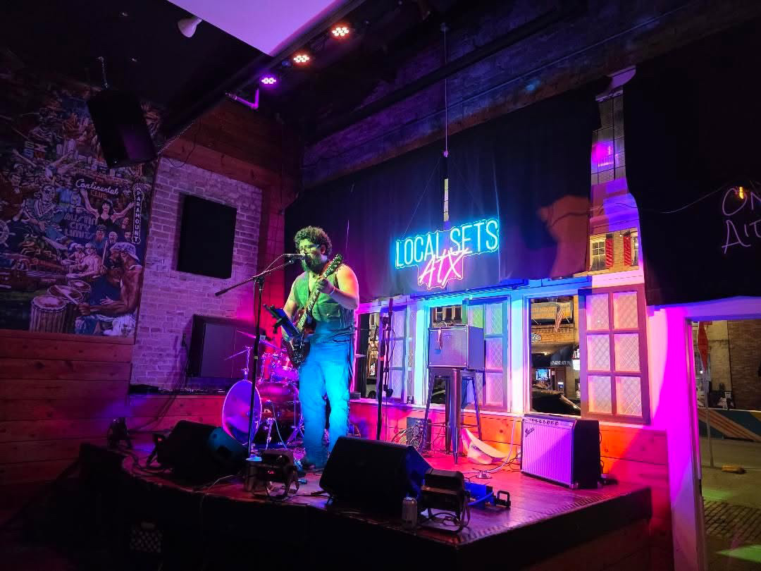 Musician performing on stage with a guitar, neon sign, and colorful set, in a cozy venue with brick walls.