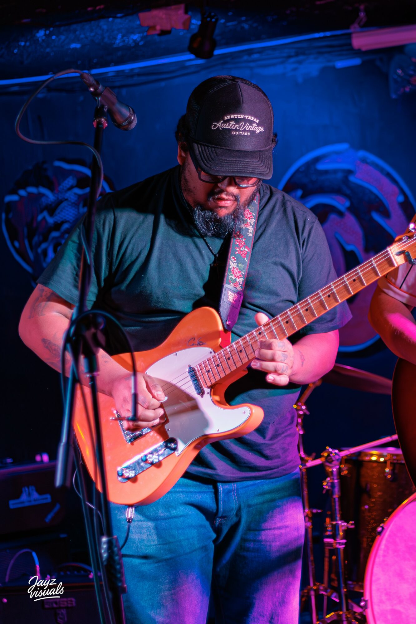 Person playing electric guitar on stage with blue and pink lighting, wearing a cap and glasses.