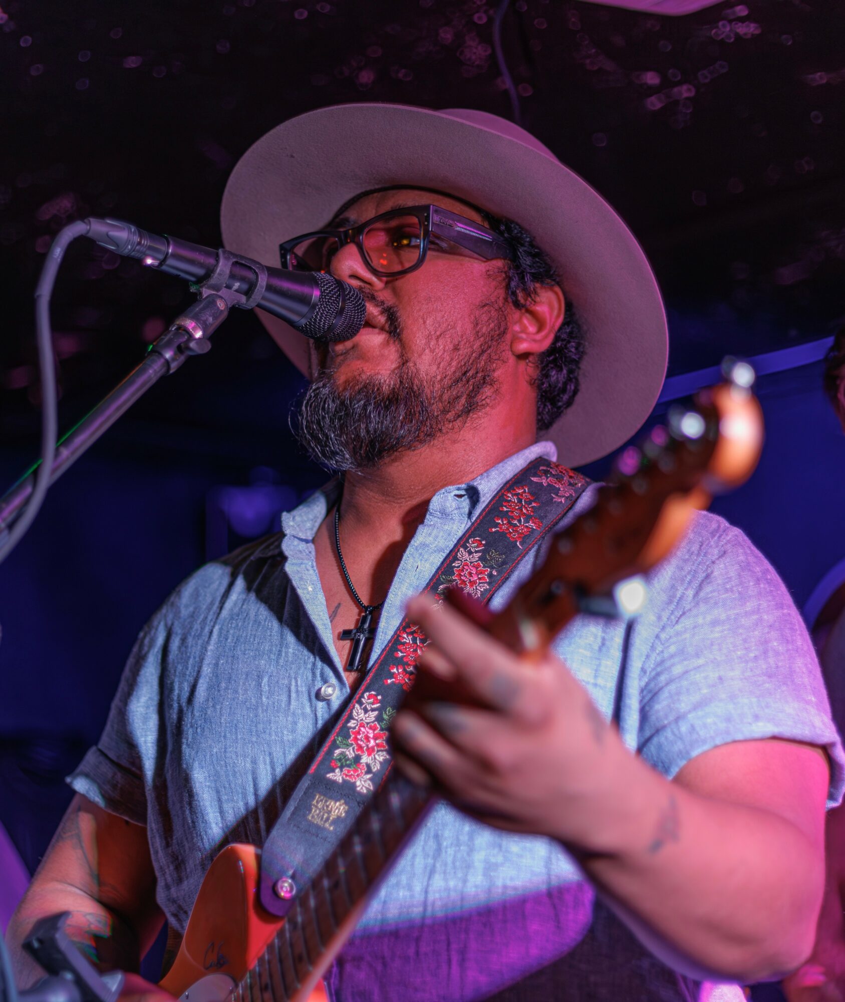 Man wearing glasses and a wide-brimmed hat plays guitar and sings into a microphone on stage.