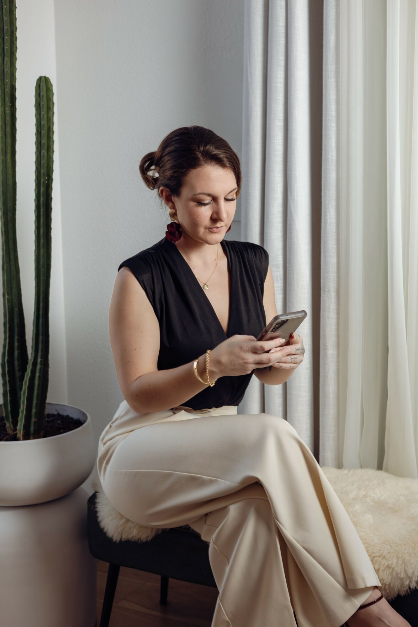 Woman sitting on a chair, looking at her phone, with a tall cactus and curtains in the background.