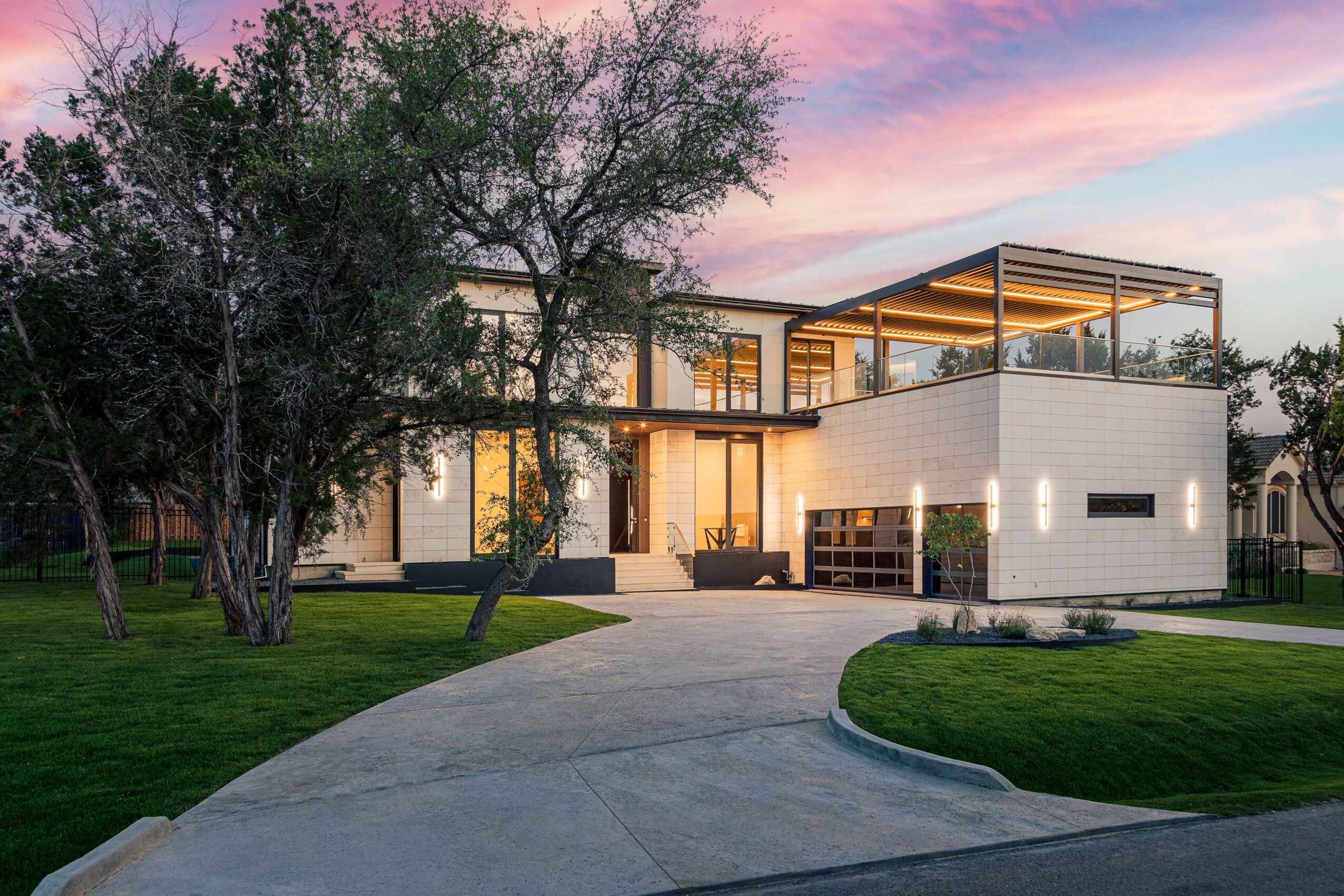 Modern house with large glass windows and a curved driveway, surrounded by trees and a green lawn, during sunset.