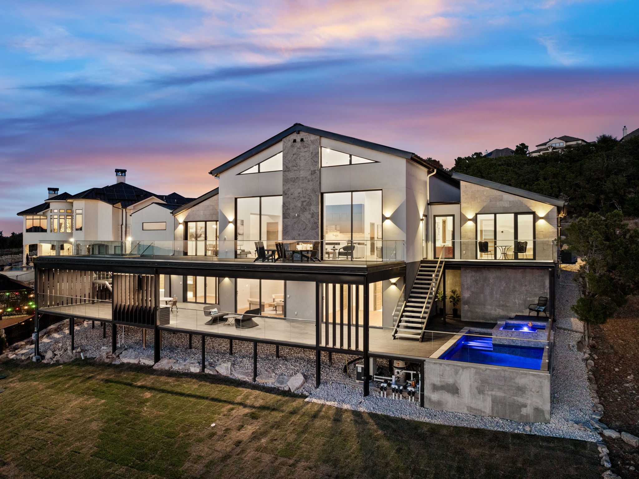 Modern house with large glass windows, outdoor deck, and pool, set against a colorful evening sky.