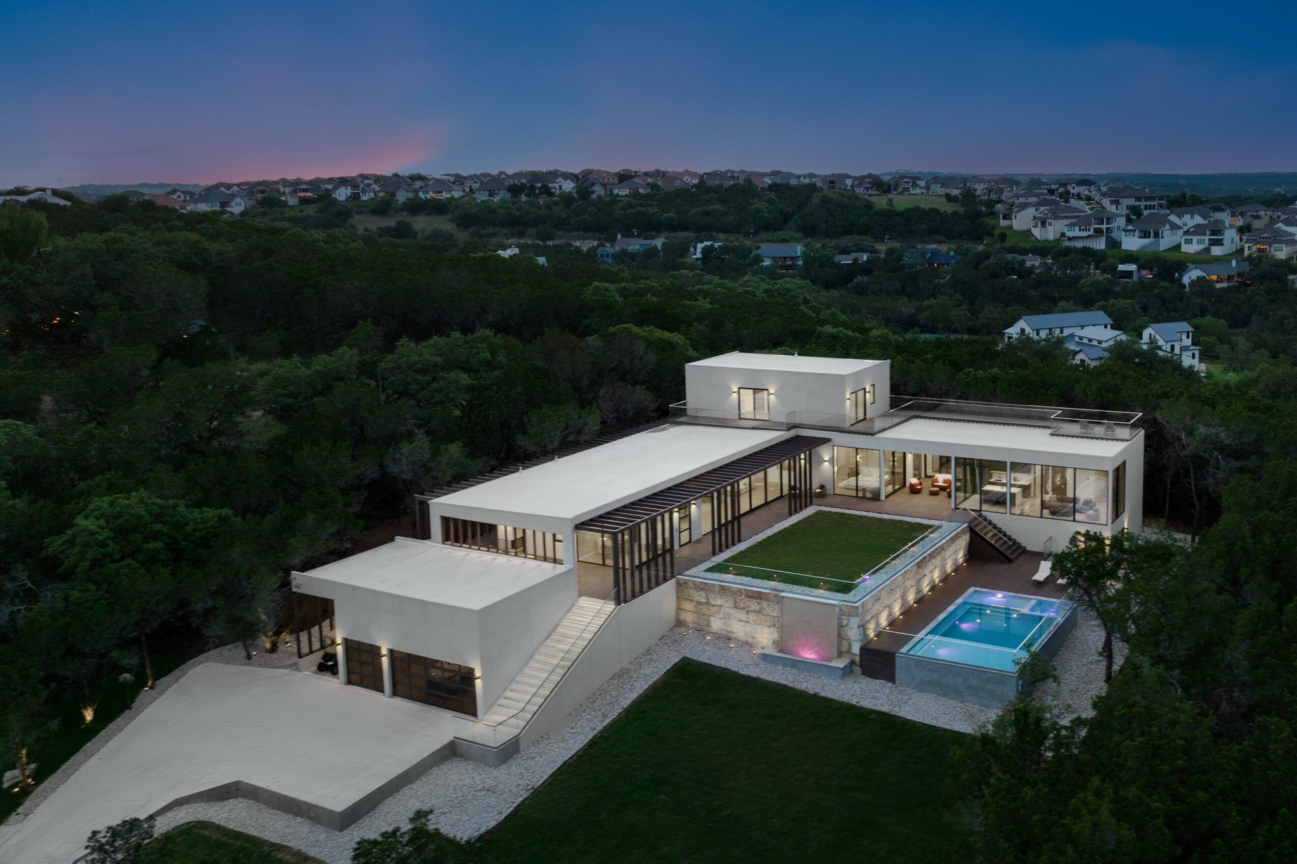 Modern house with pool and green lawn, surrounded by trees, under a twilight sky.