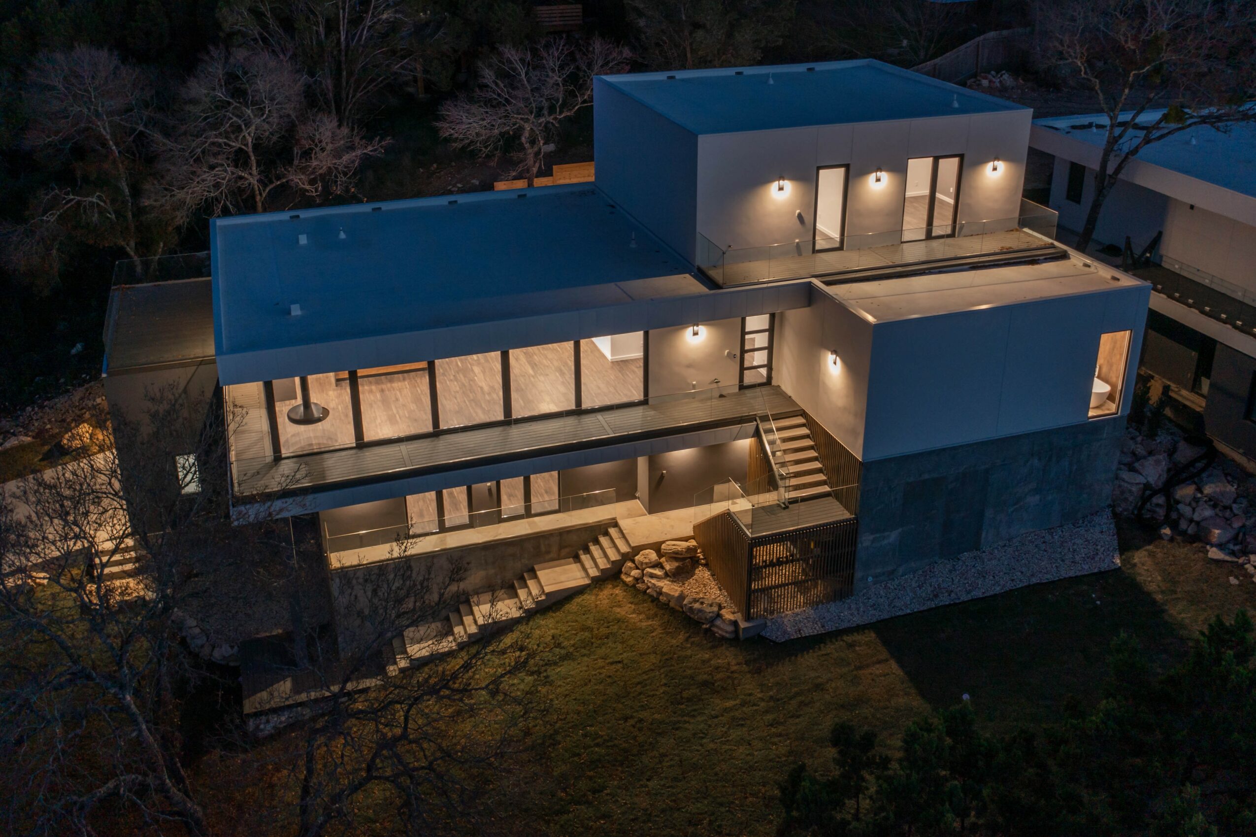 Modern multi-level house with large glass windows, illuminated exterior, and outdoor stairs, surrounded by trees and grass at night.
