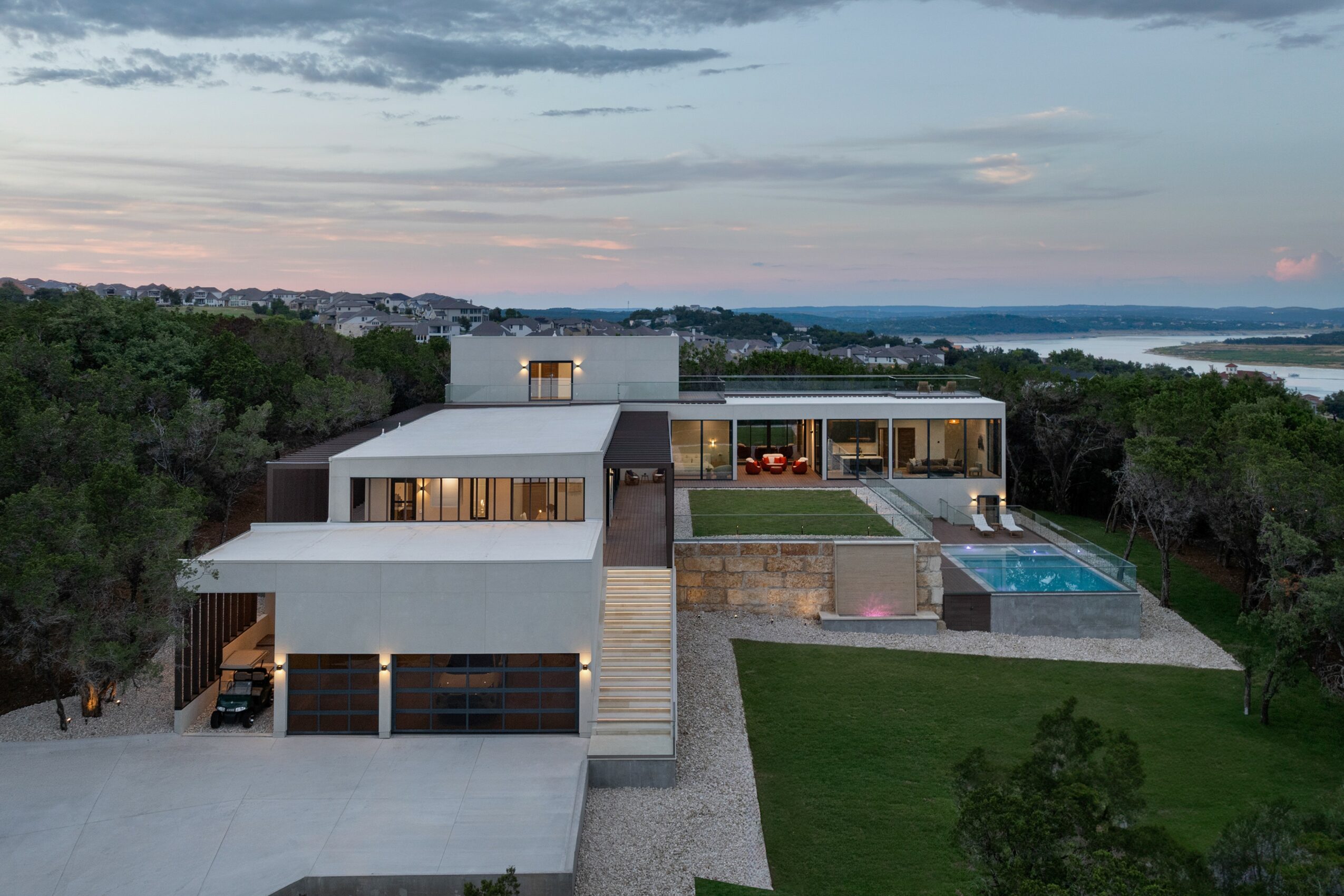 Modern house with outdoor pool, large windows, and multiple levels, surrounded by greenery and overlooking a body of water.