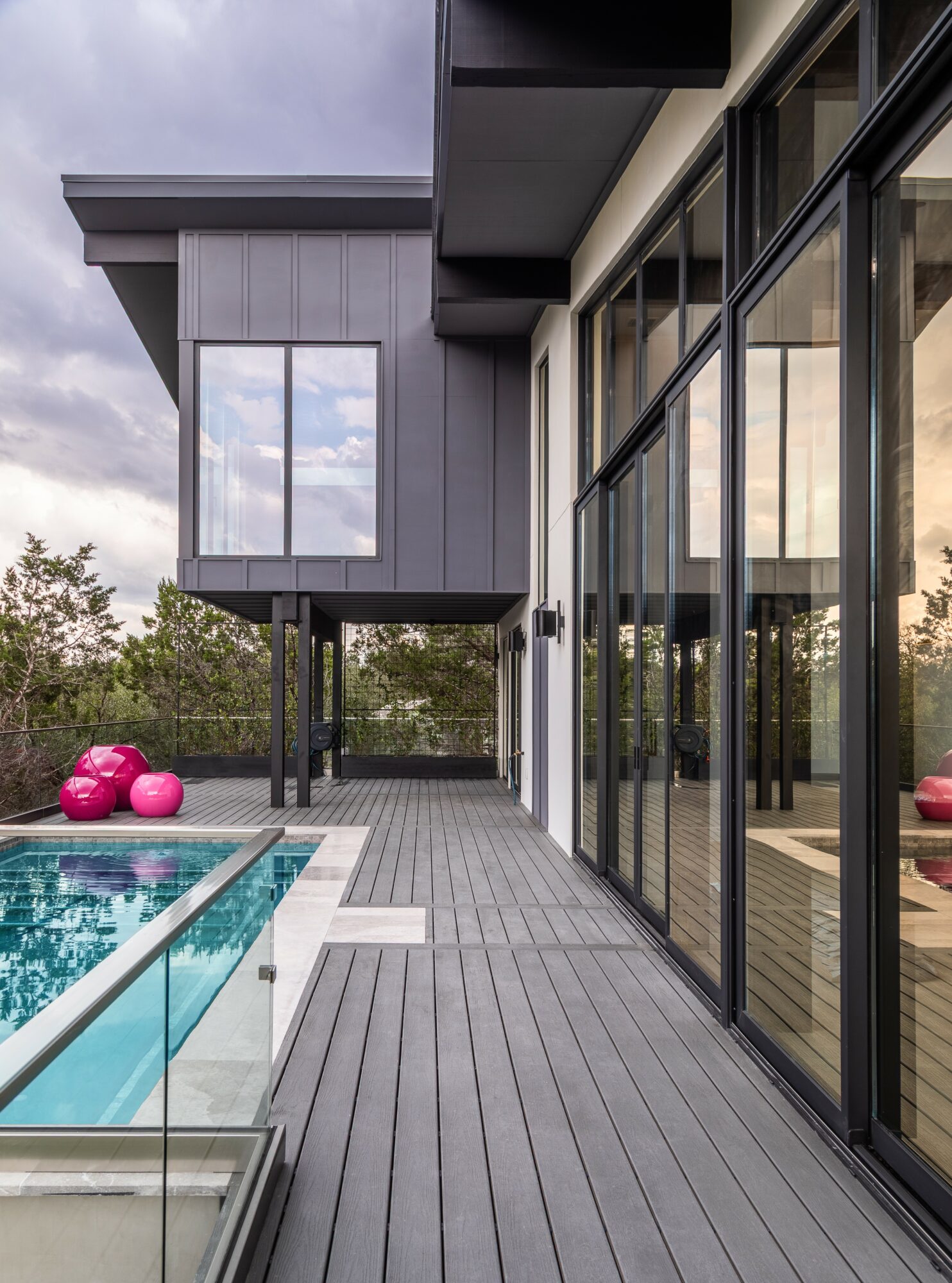 Modern house exterior with large glass windows, a wooden deck, and a swimming pool, under a cloudy sky.