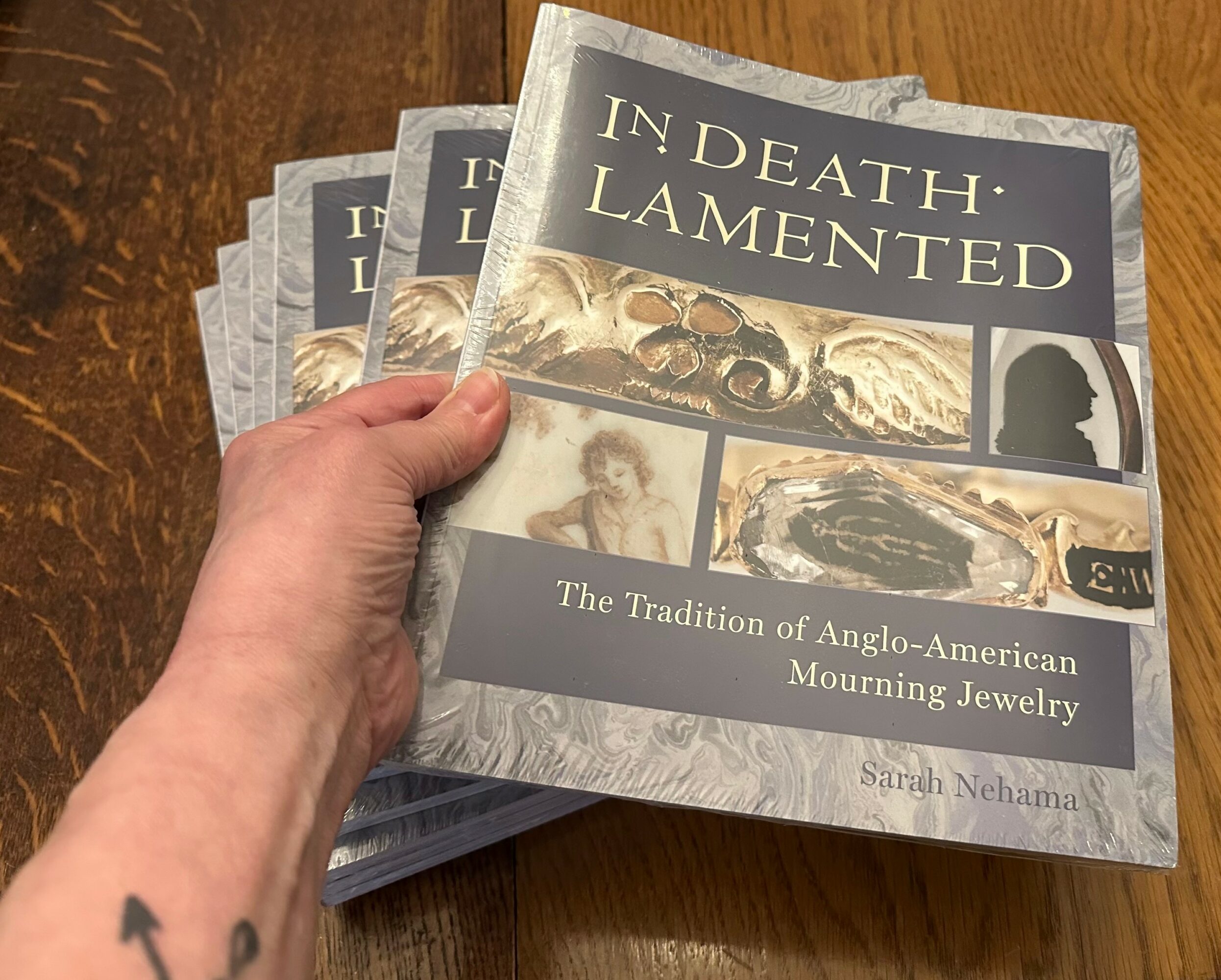 Hand holding multiple copies of a book titled 'In Death Lamented' about Anglo-American mourning jewelry, on a wooden surface.