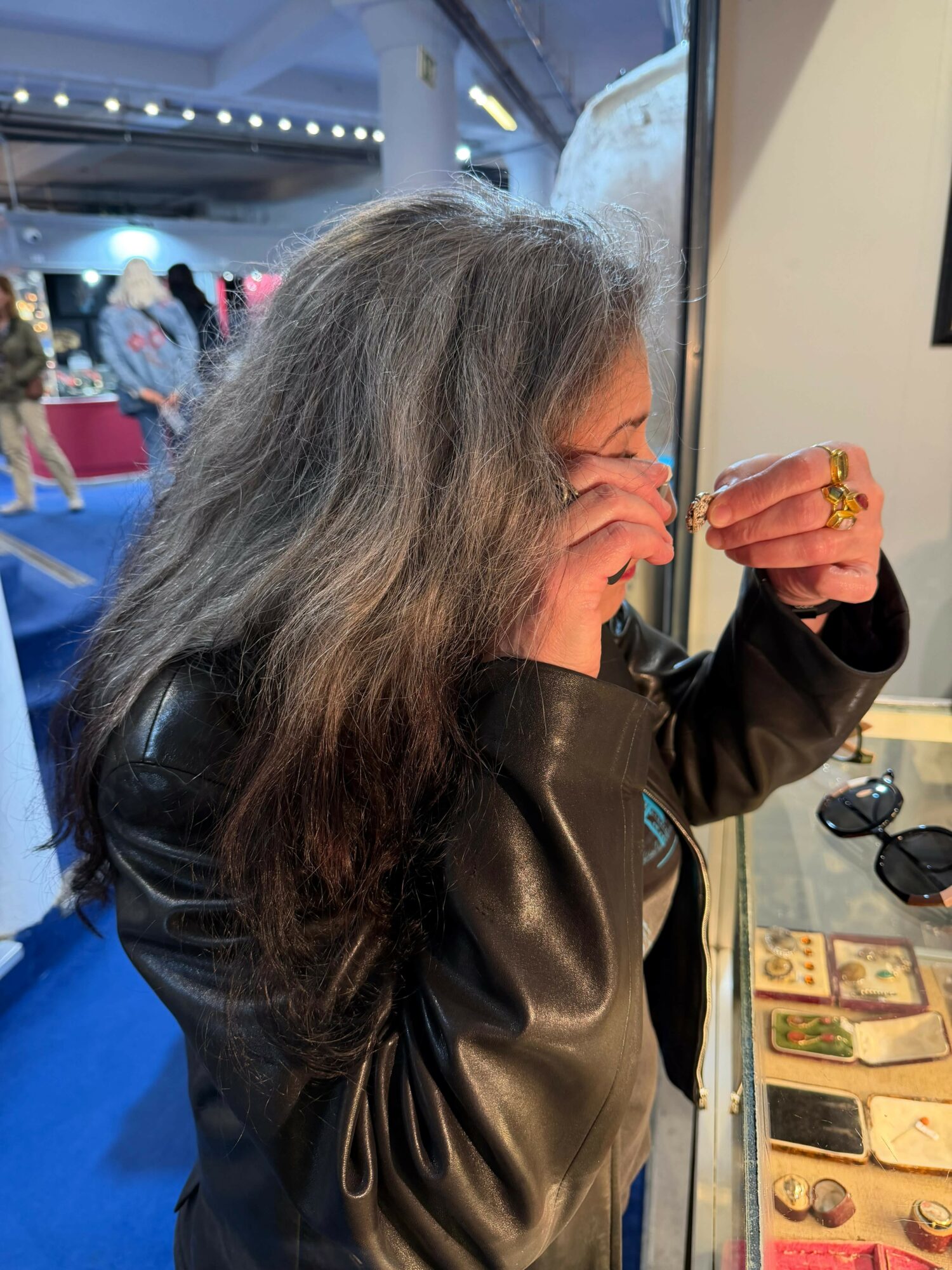 Woman with long, wavy hair wearing a black leather jacket, covering her face with her hand, in an indoor setting.