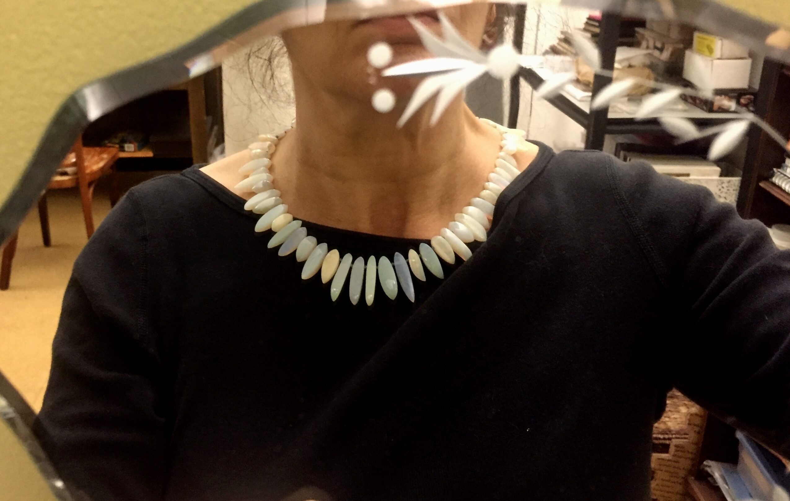 Person wearing a black top and a white beaded necklace, taking a mirror selfie in a room with books and furniture