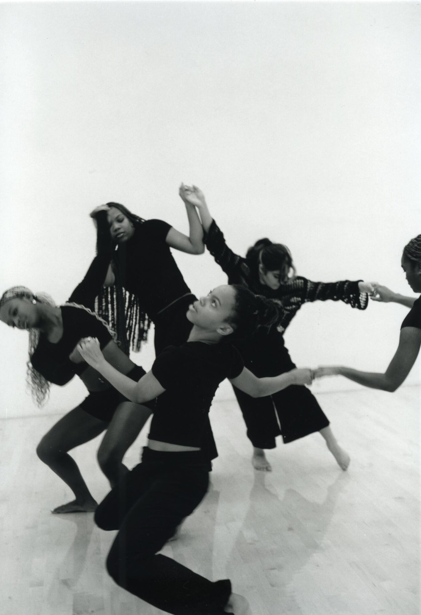 Group of five people holding hands and leaning back in a dance or performance, with a plain background.