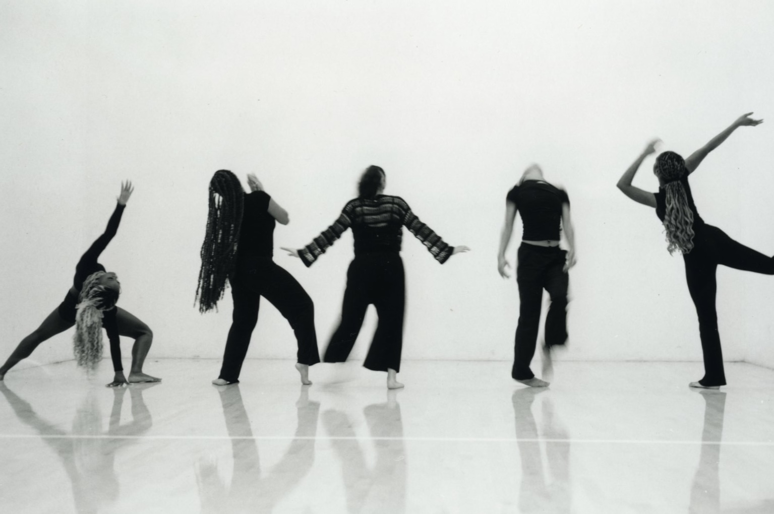 Five people in dance poses against a plain background, some with arms raised or extended, in black clothing.