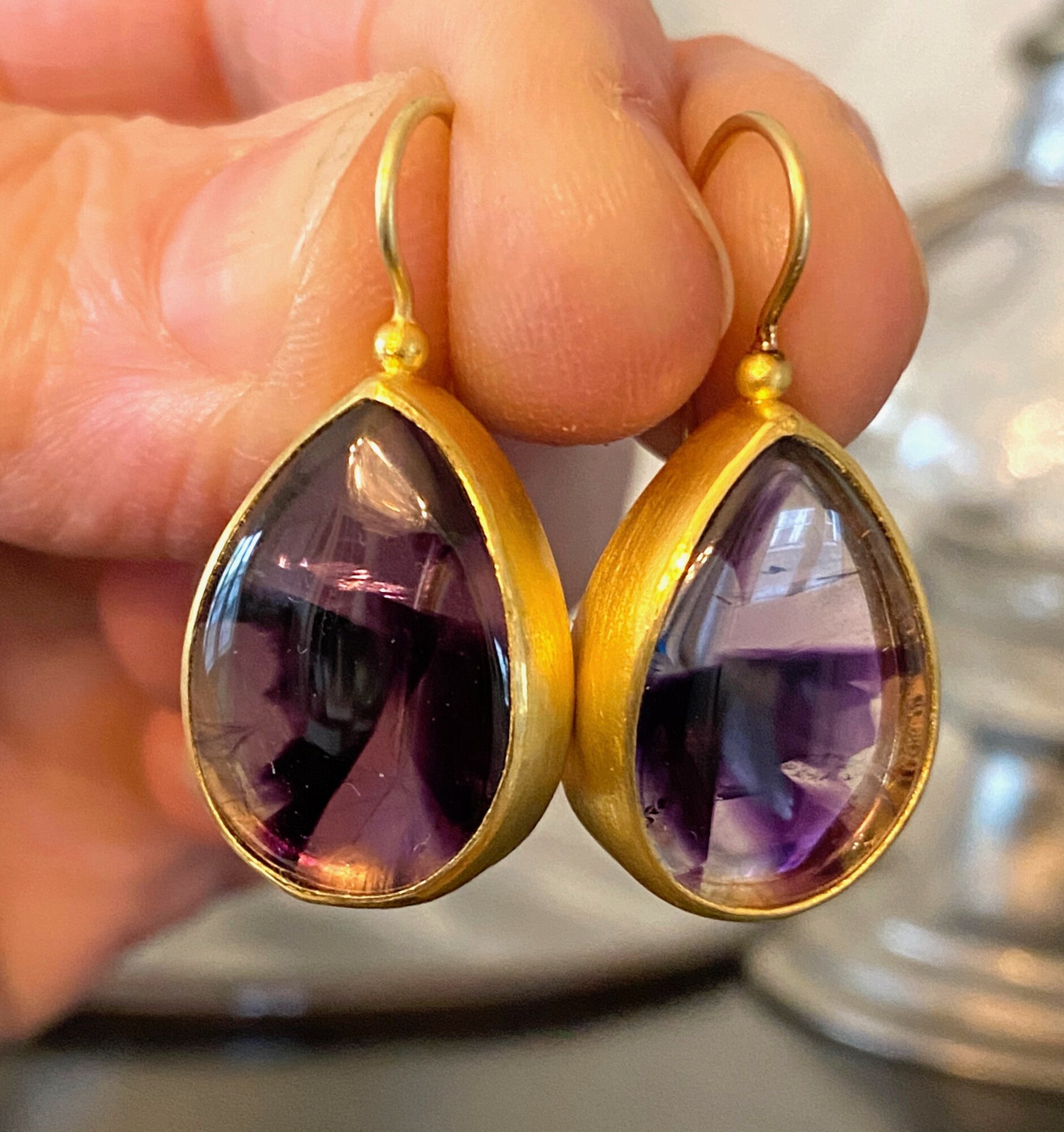 Gold teardrop-shaped earrings with purple gemstones held by a hand.