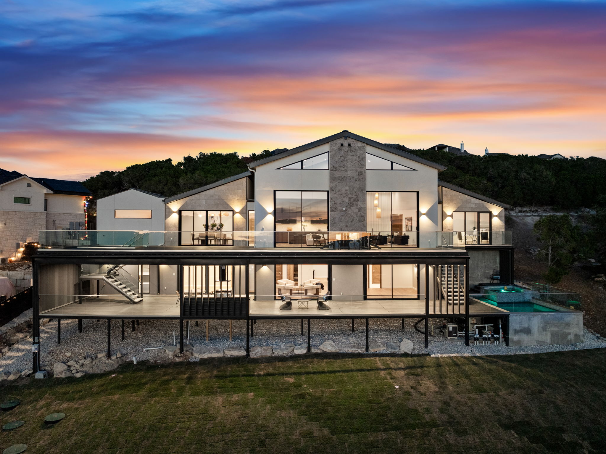 Modern multi-story house with large windows, balcony, and pool at sunset, overlooking a grassy area and hills.