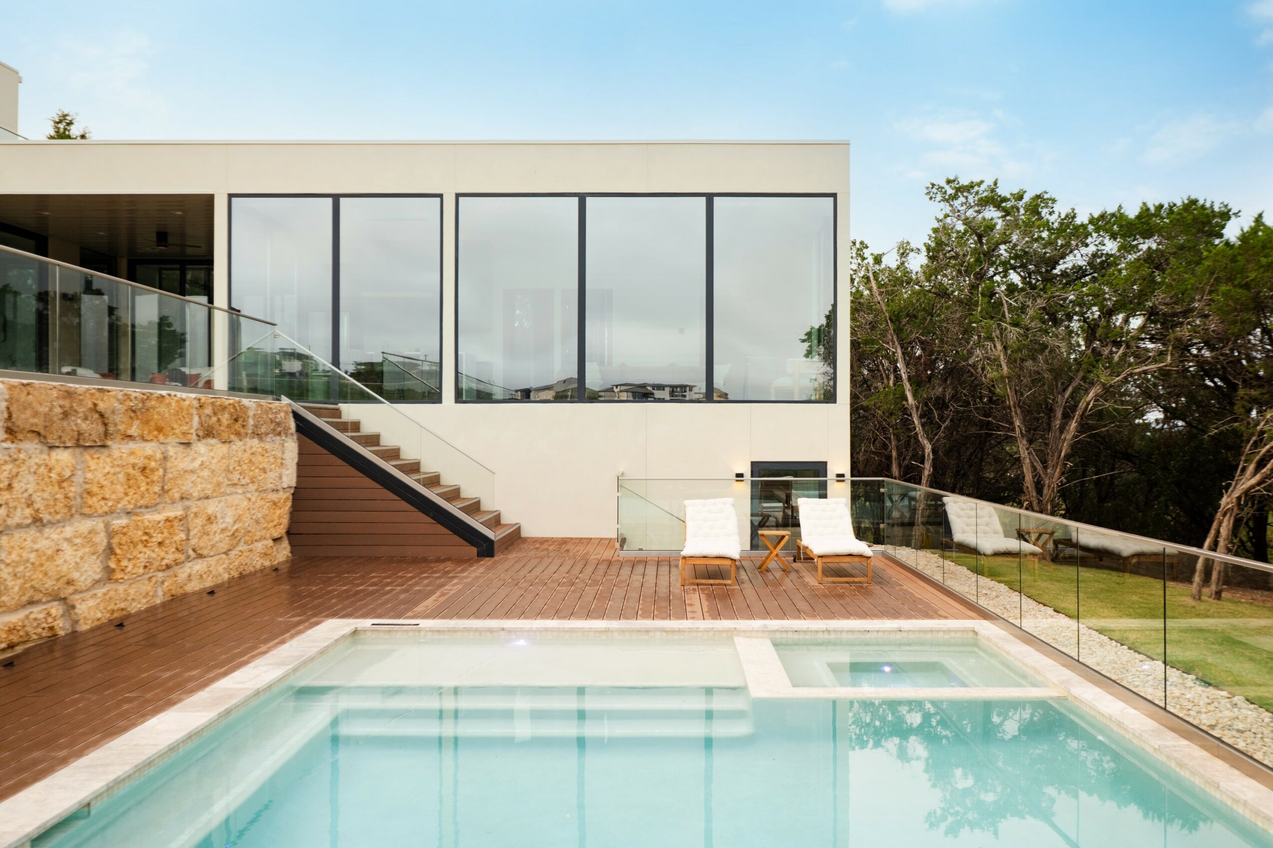Modern house with large glass windows, outdoor pool, wooden deck, and glass railing, surrounded by trees and blue sky.