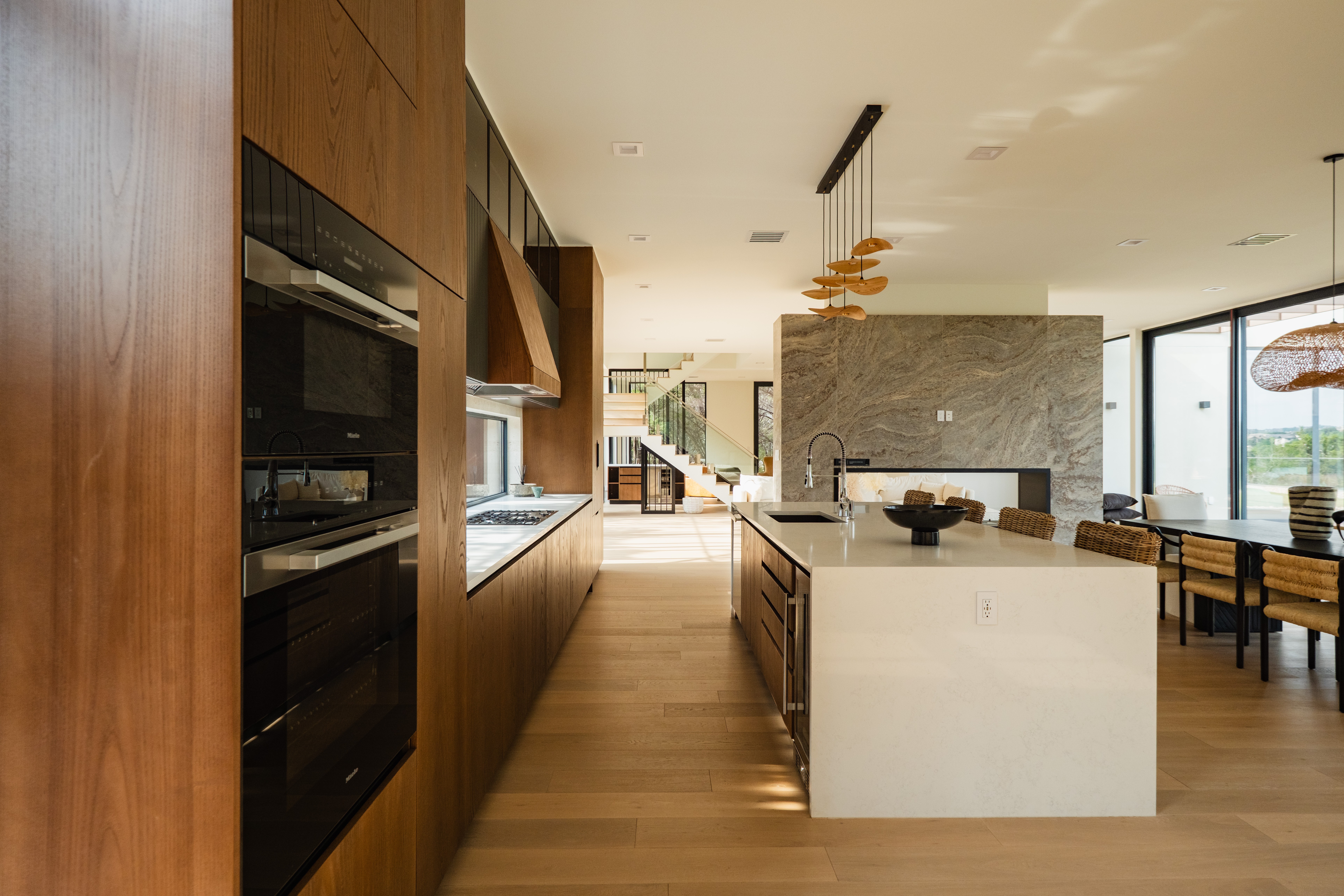 Modern kitchen with wooden cabinets, white island, and open living area with large windows and dining table.