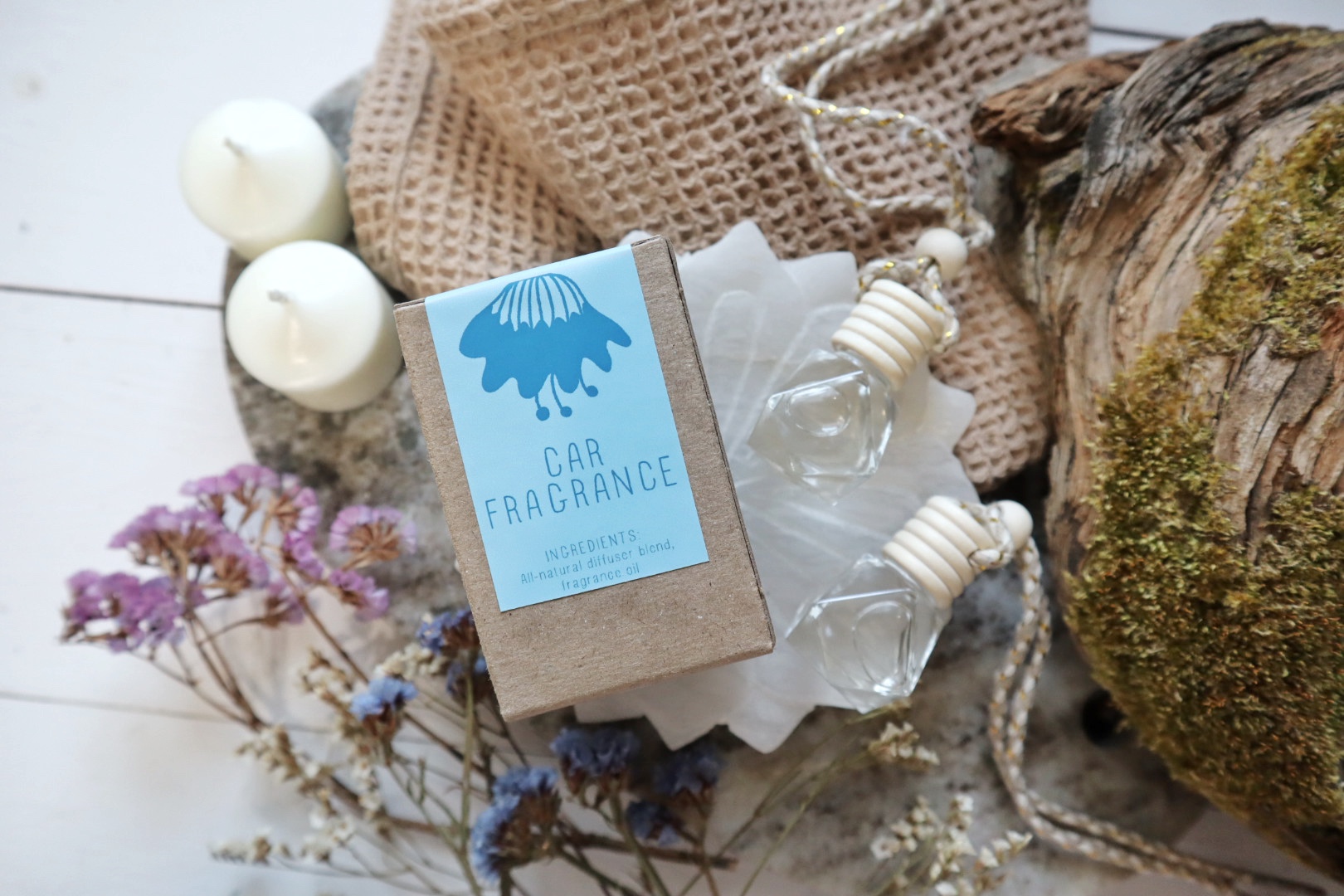 A cardboard box labeled 'Car Fragrance' surrounded by candles, a glass bottle, dried flowers, a burlap cloth, and a tree trunk.