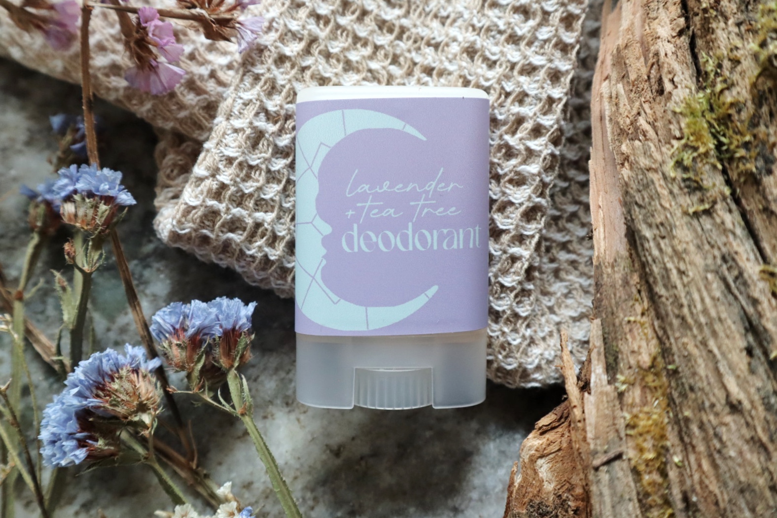 A deodorant stick with a pastel purple and white label, placed on a textured surface with flowers and wood nearby.