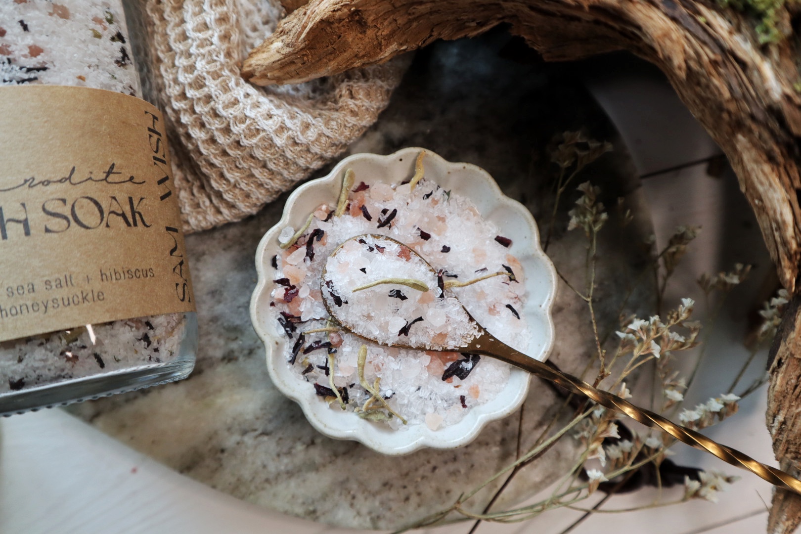 Small decorative dish with dried flower petals and a gold spoon, surrounded by natural elements and a jar with a label.