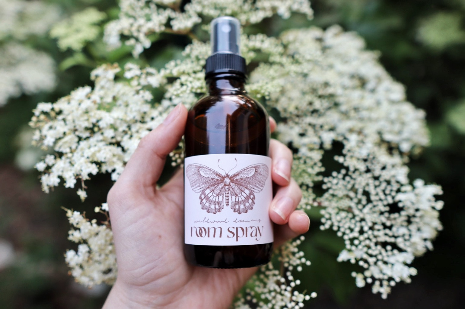 Hand holding a brown spray bottle with a white label and black spray nozzle, in front of white flowering plant