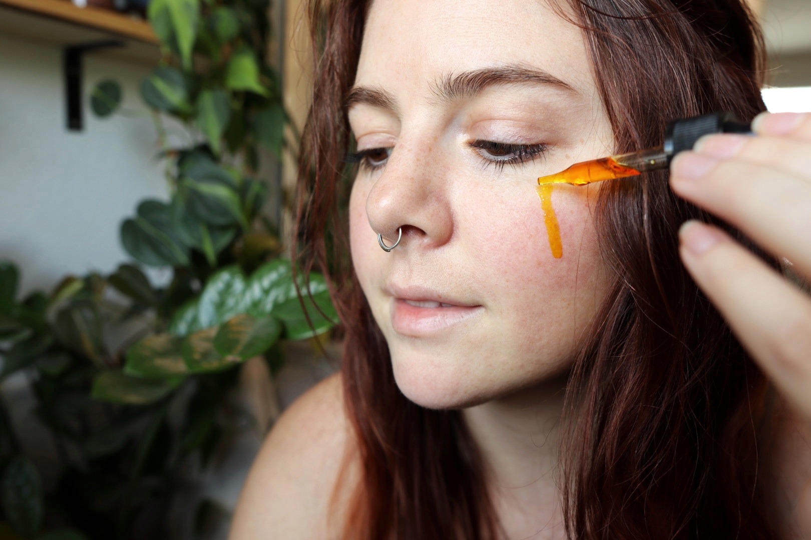 Person with red hair and nose piercing applying orange liquid to face with a dropper, indoors with green plants in background.