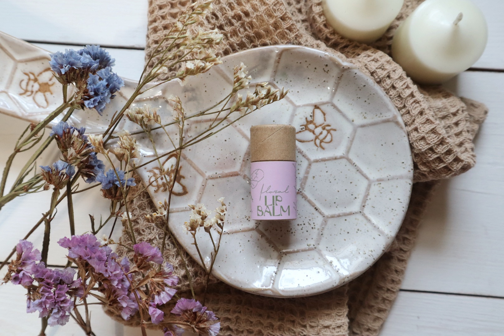 Decorative plate with a small bottle, dried flowers, candles, and textured cloth on a white surface.