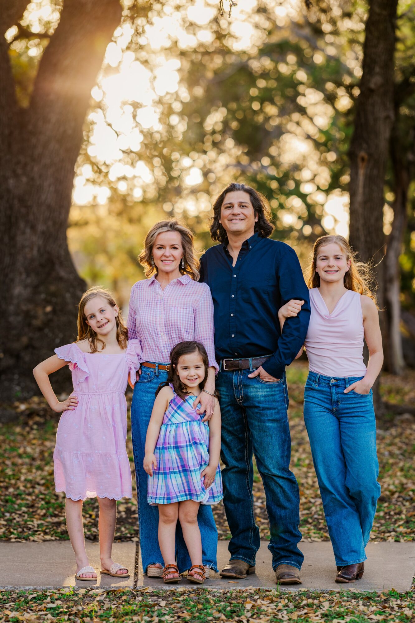 Family of five standing outdoors in a park with trees and sunlight, smiling, with three girls and one man and woman.