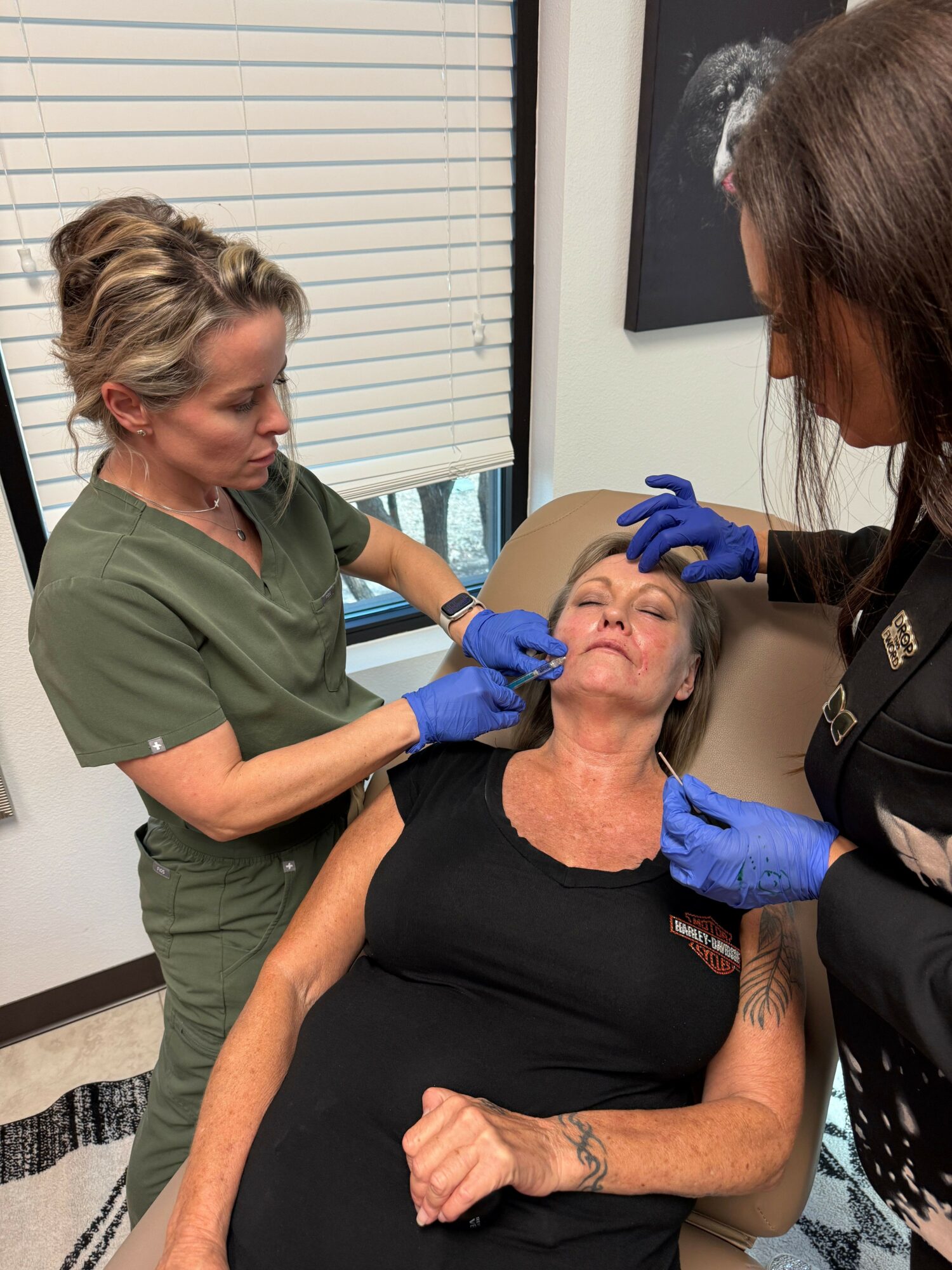 A woman in green scrubs administers a nasal swab to a woman in black sitting in a chair, with a healthcare worker wearing gloves.