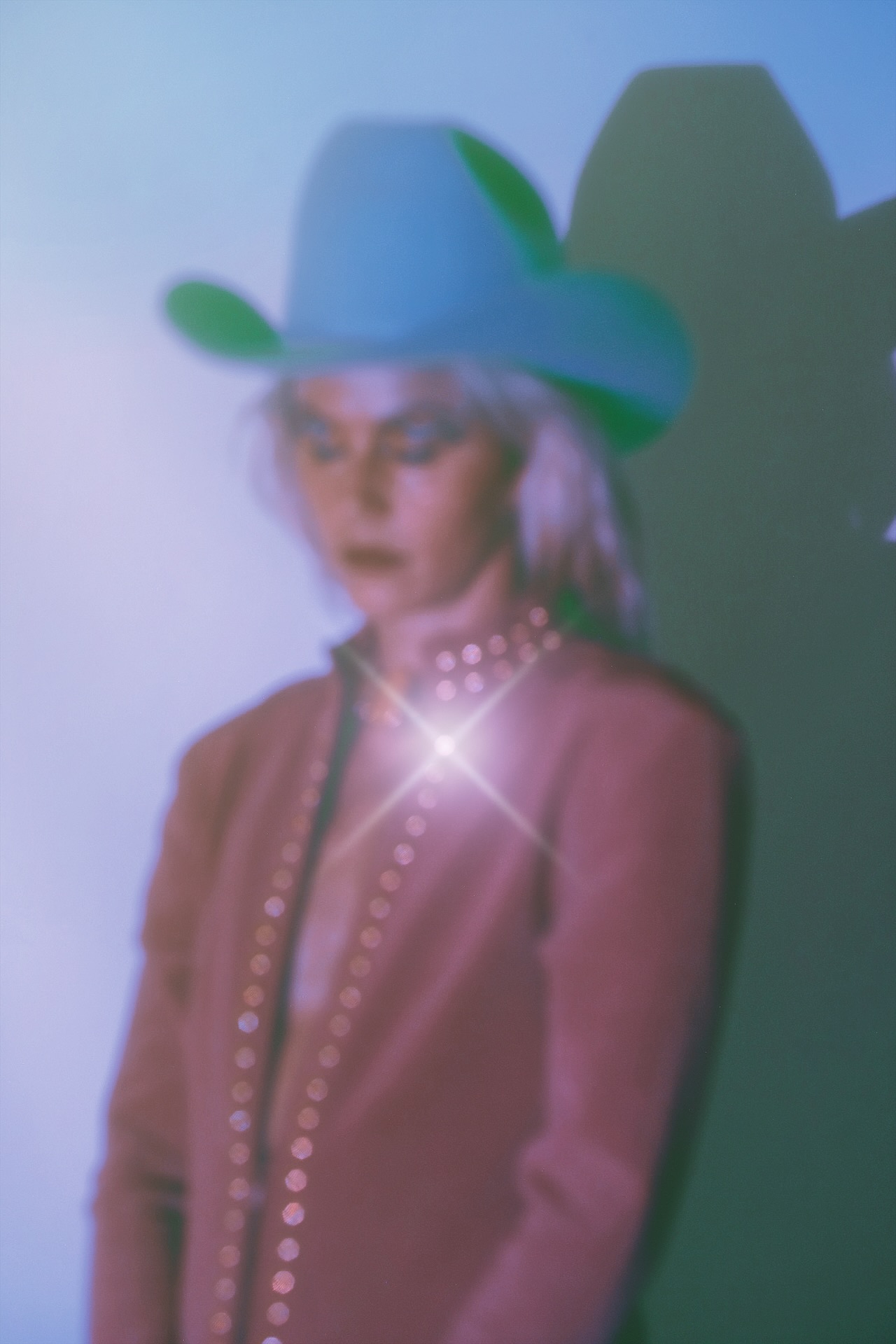 Person wearing a large hat with a wide brim, pearl necklace, and pink jacket, standing against a dark background.