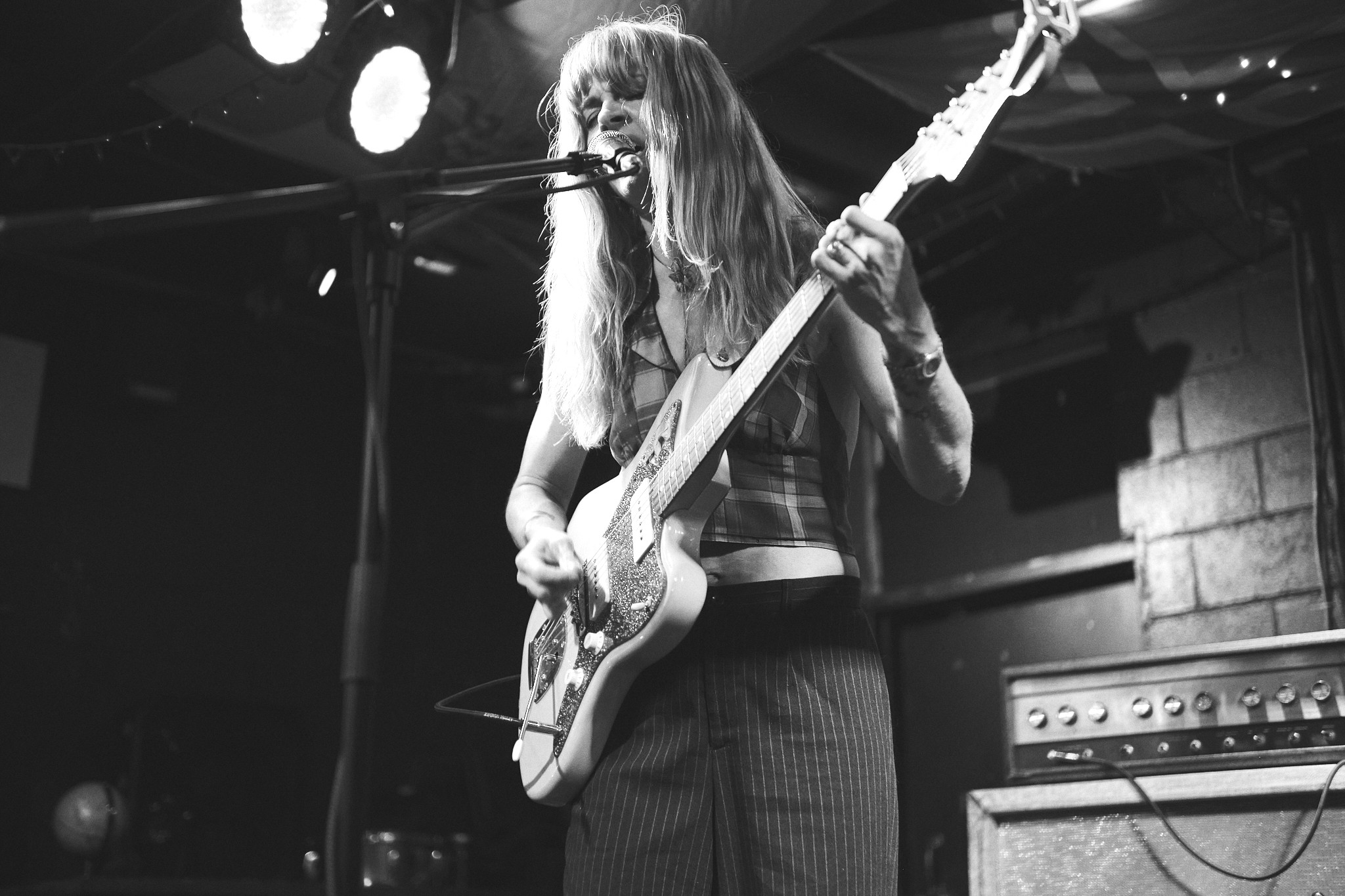 Female musician playing guitar and singing into microphone on stage, with amplifier behind her, in black and white.