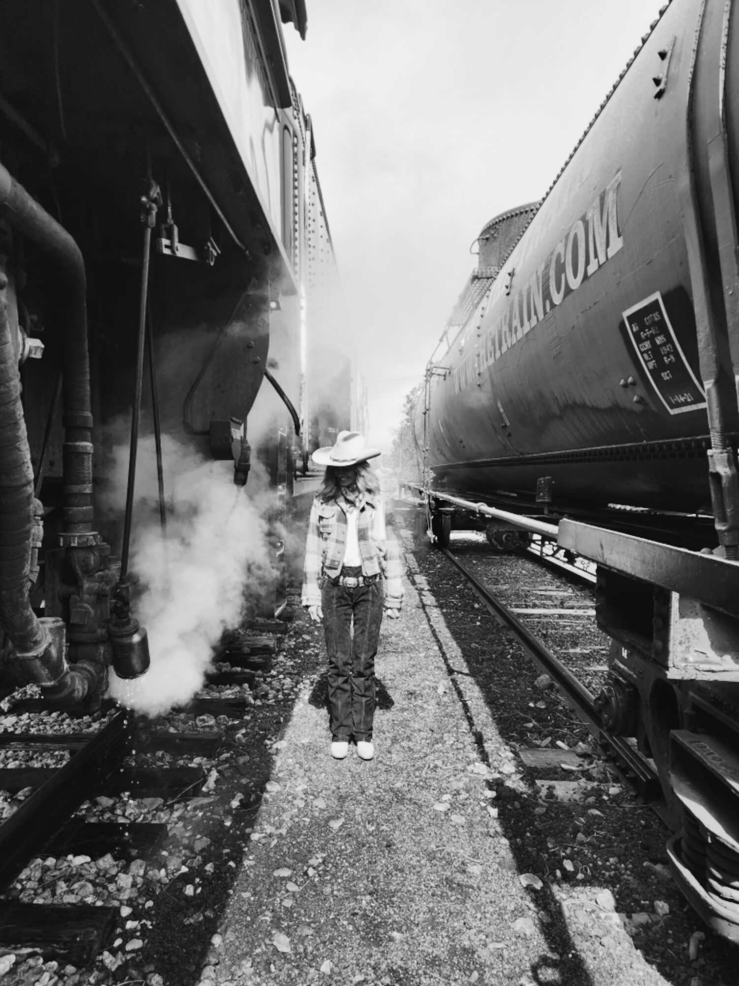 Child standing between two trains, one emitting steam, on railway tracks, wearing a hat and casual clothing.