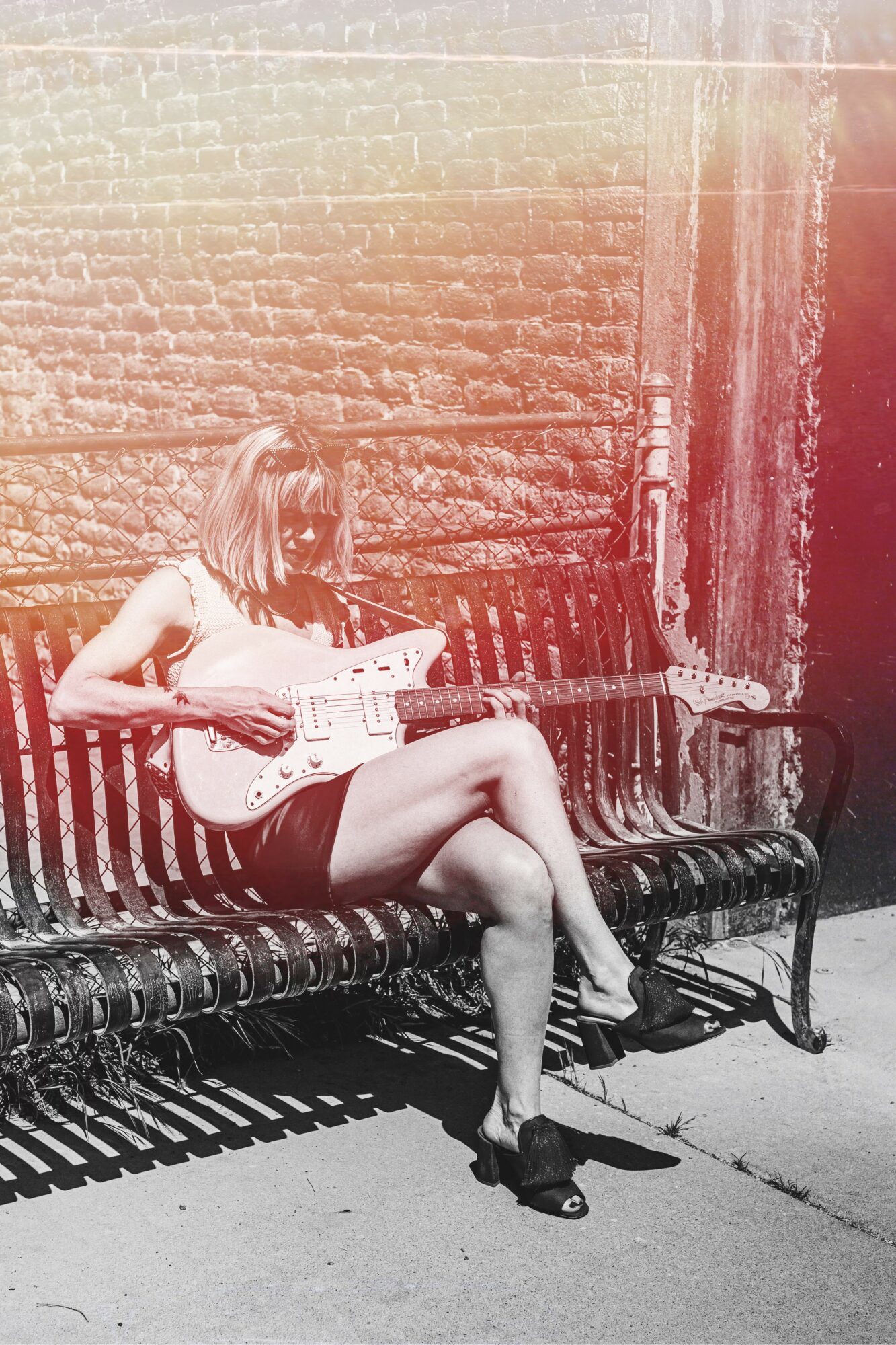 Woman sitting on a bench playing an electric guitar outdoors, with a brick wall behind her.