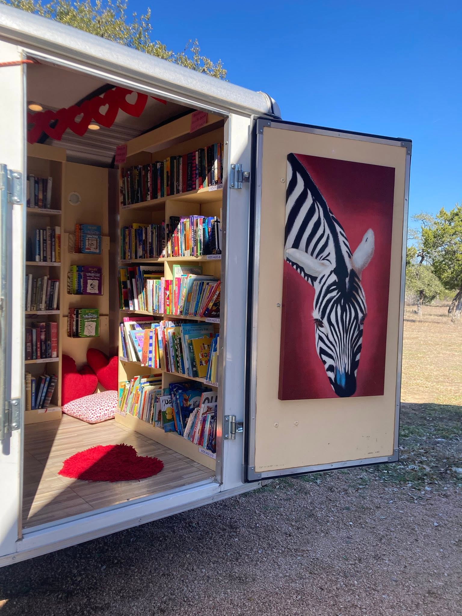 Open mobile library with bookshelves, a zebra painting on the door, and a red rug inside, outdoors on a sunny day.