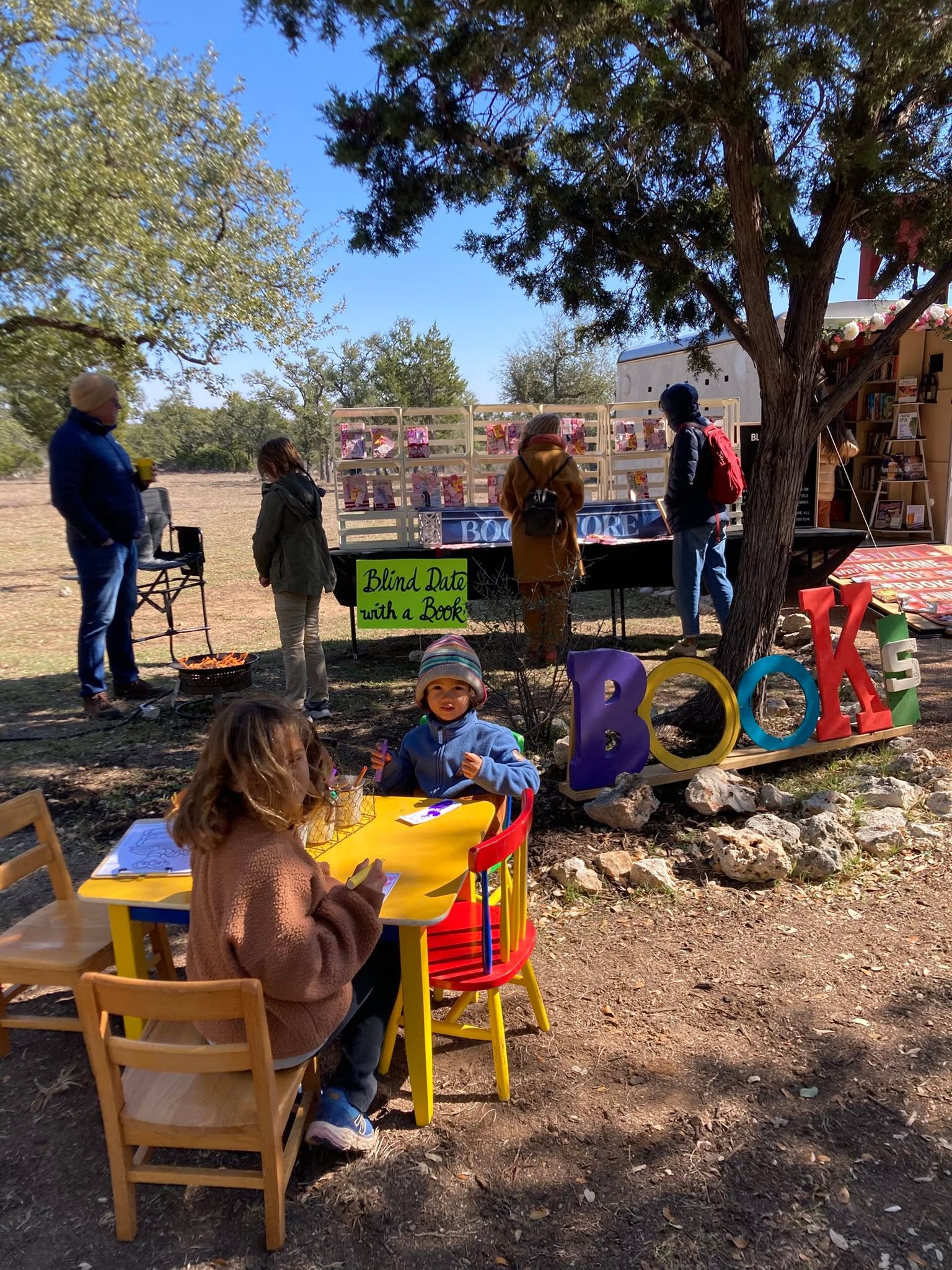 Children sit at a yellow table outdoors, with colorful letters spelling 'BOOKS' nearby, and people browsing a market stall under trees.