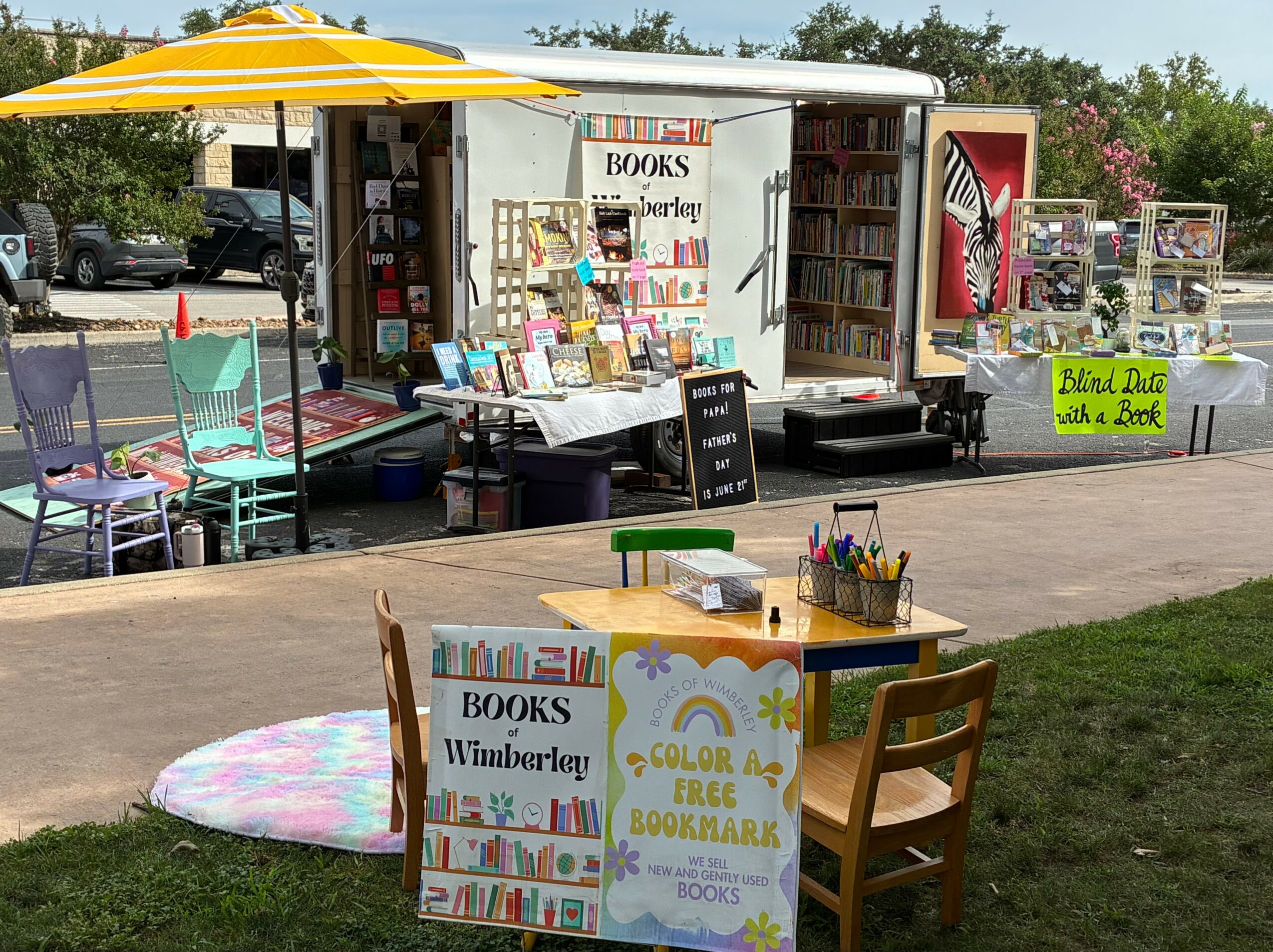 Mobile book stand with books, chairs, and a yellow umbrella outdoors, with a sidewalk and trees in background.