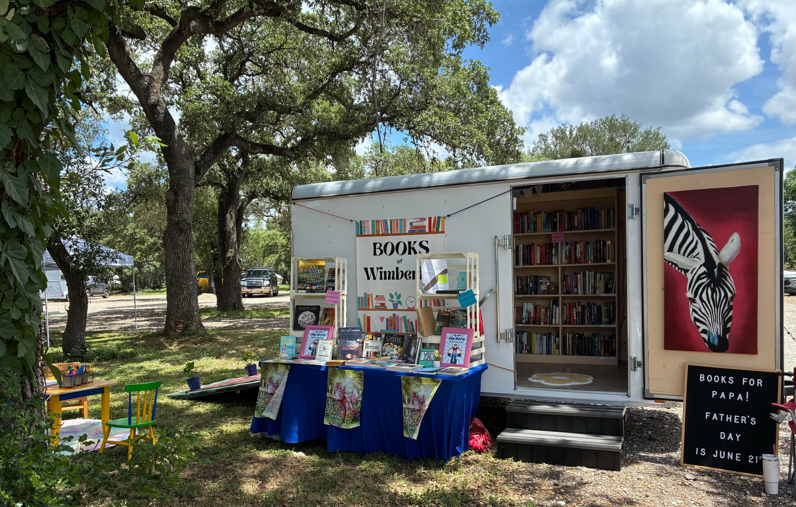 Mobile book trailer with books displayed outside, surrounded by trees and a grassy area, under a partly cloudy sky.