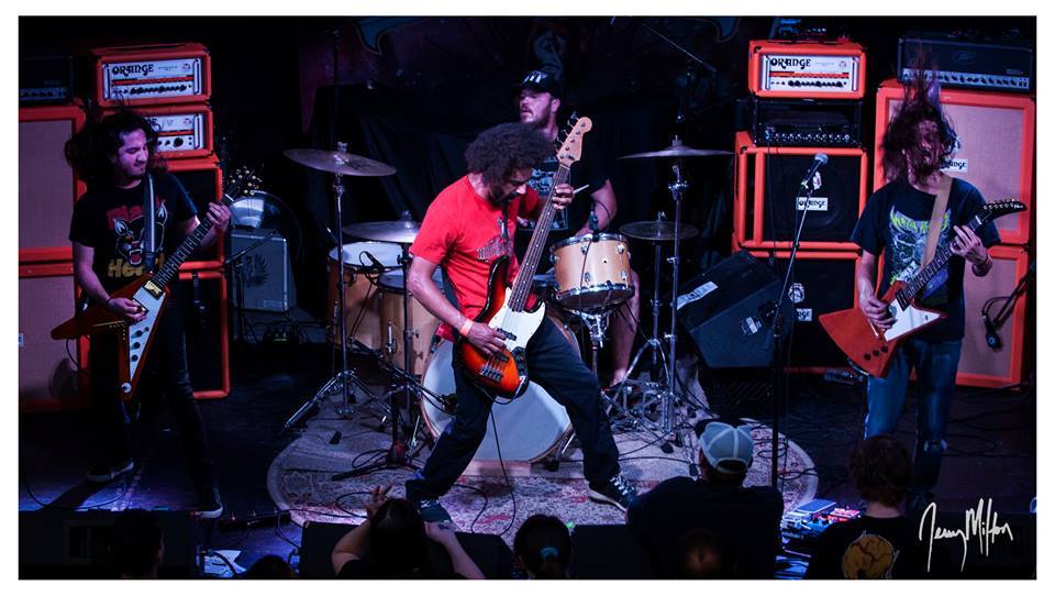 Band performing on stage with guitars, drums, and orange amplifiers, dark background, audience visible at bottom, lively scene.