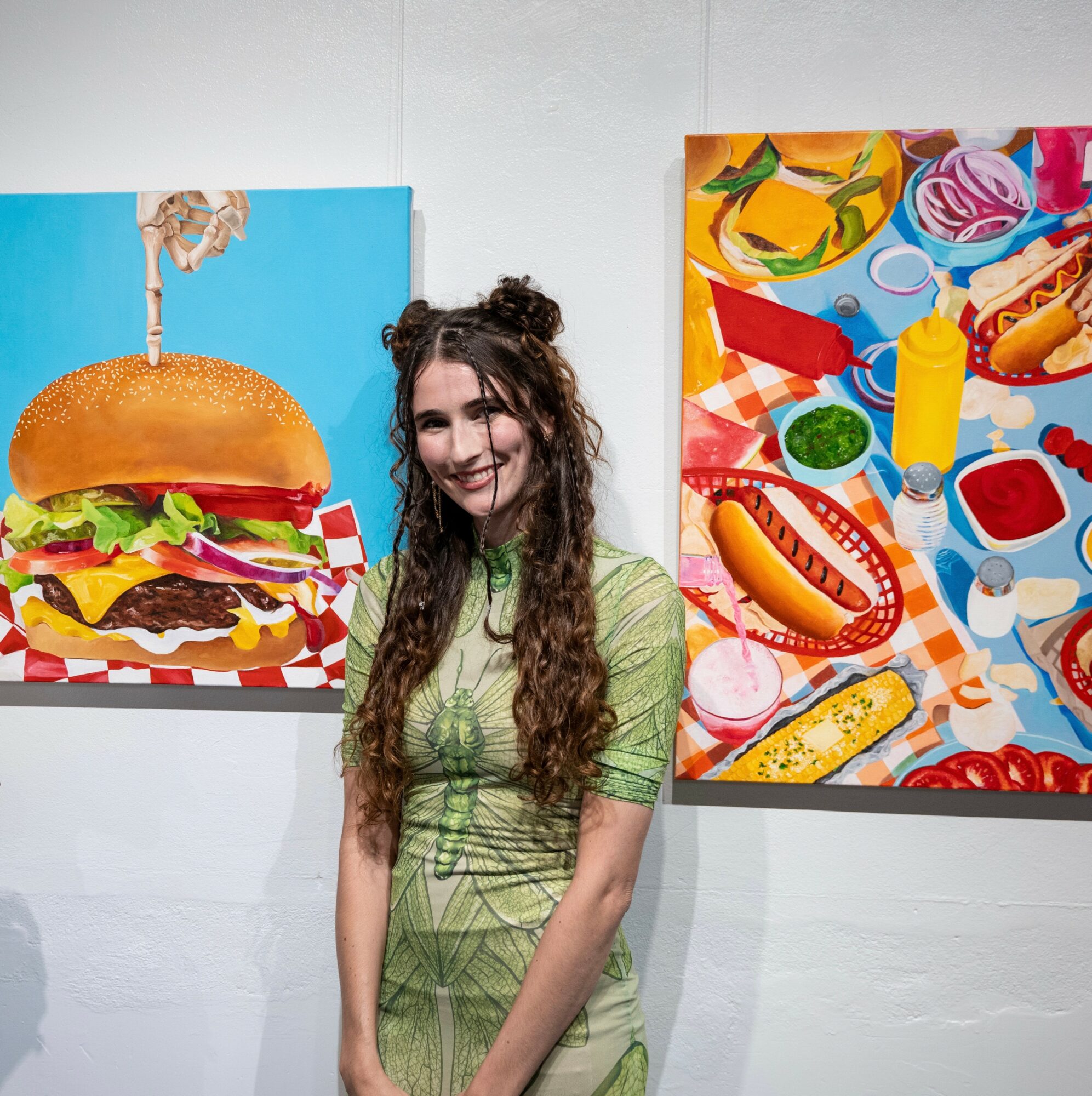 Young woman with long curly hair smiling, standing between colorful food-themed paintings on a white wall.