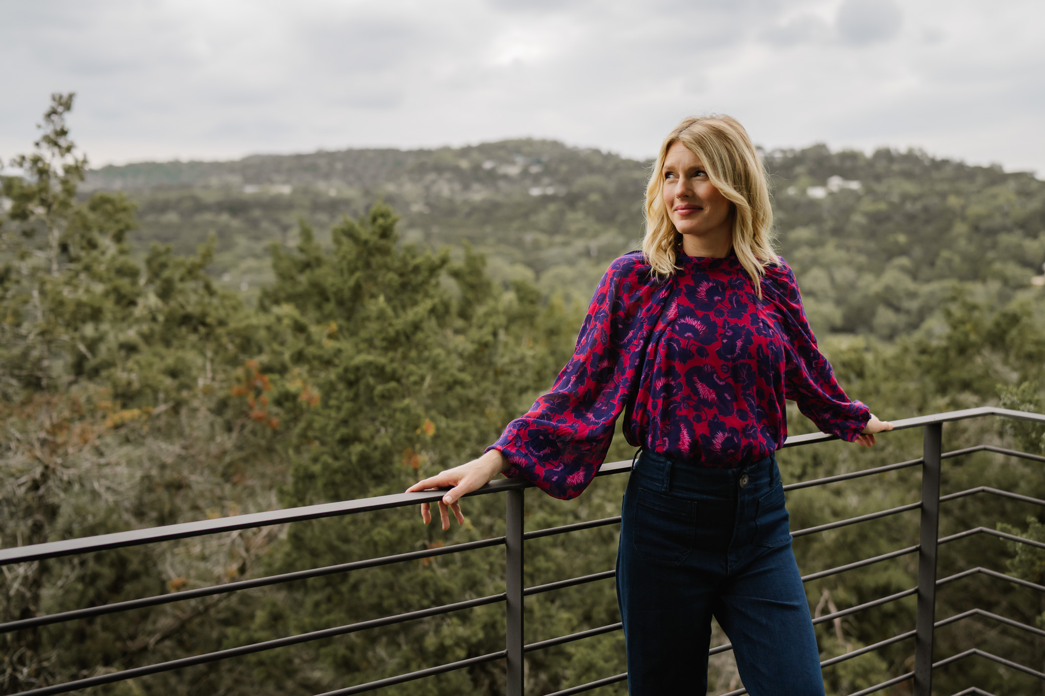 Woman with blonde hair wearing a purple patterned blouse and dark pants standing outdoors with a railing and trees in the background.