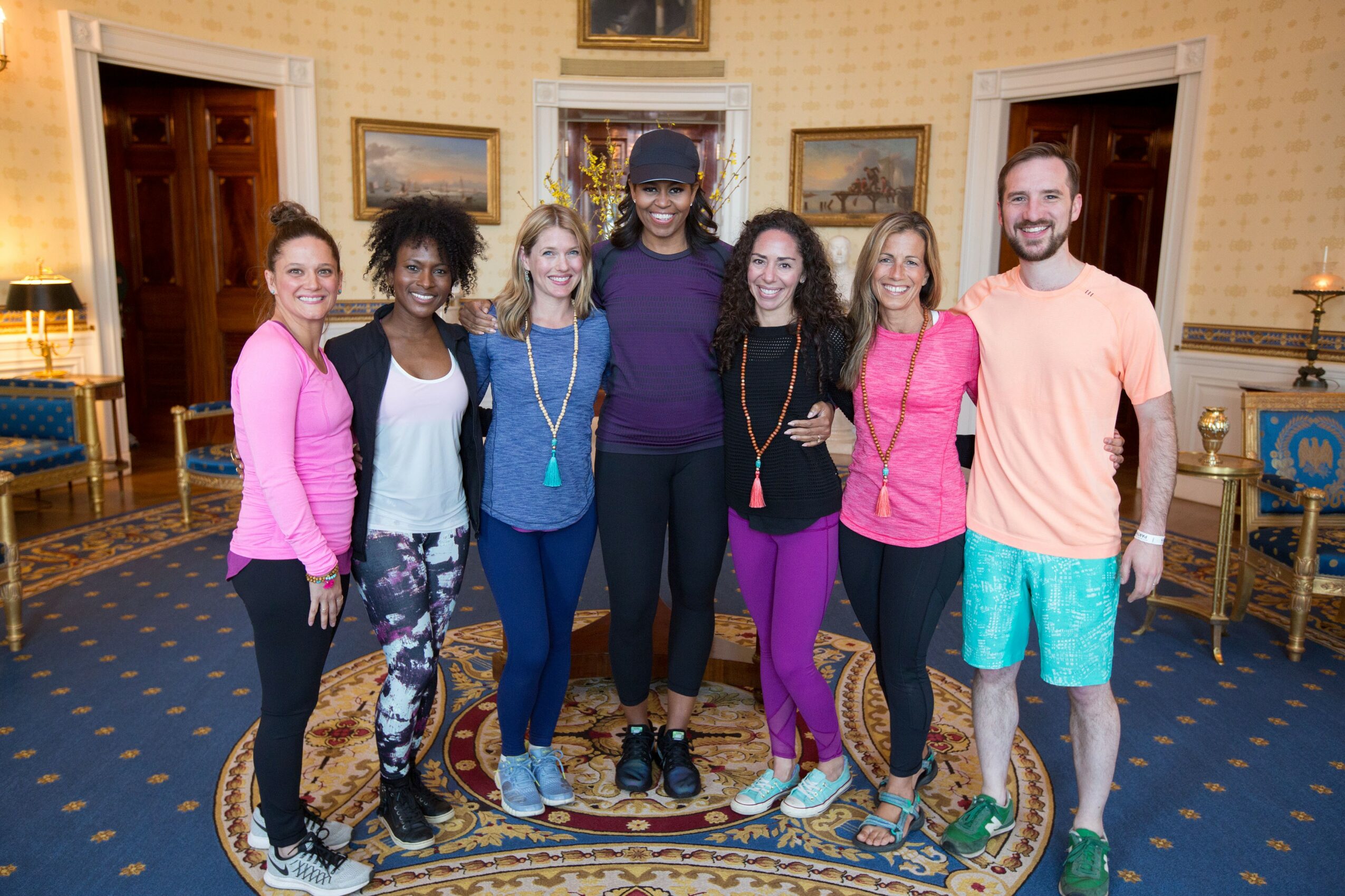Group of seven people standing together in a decorated room with yellow walls and blue carpet, smiling at the camera.