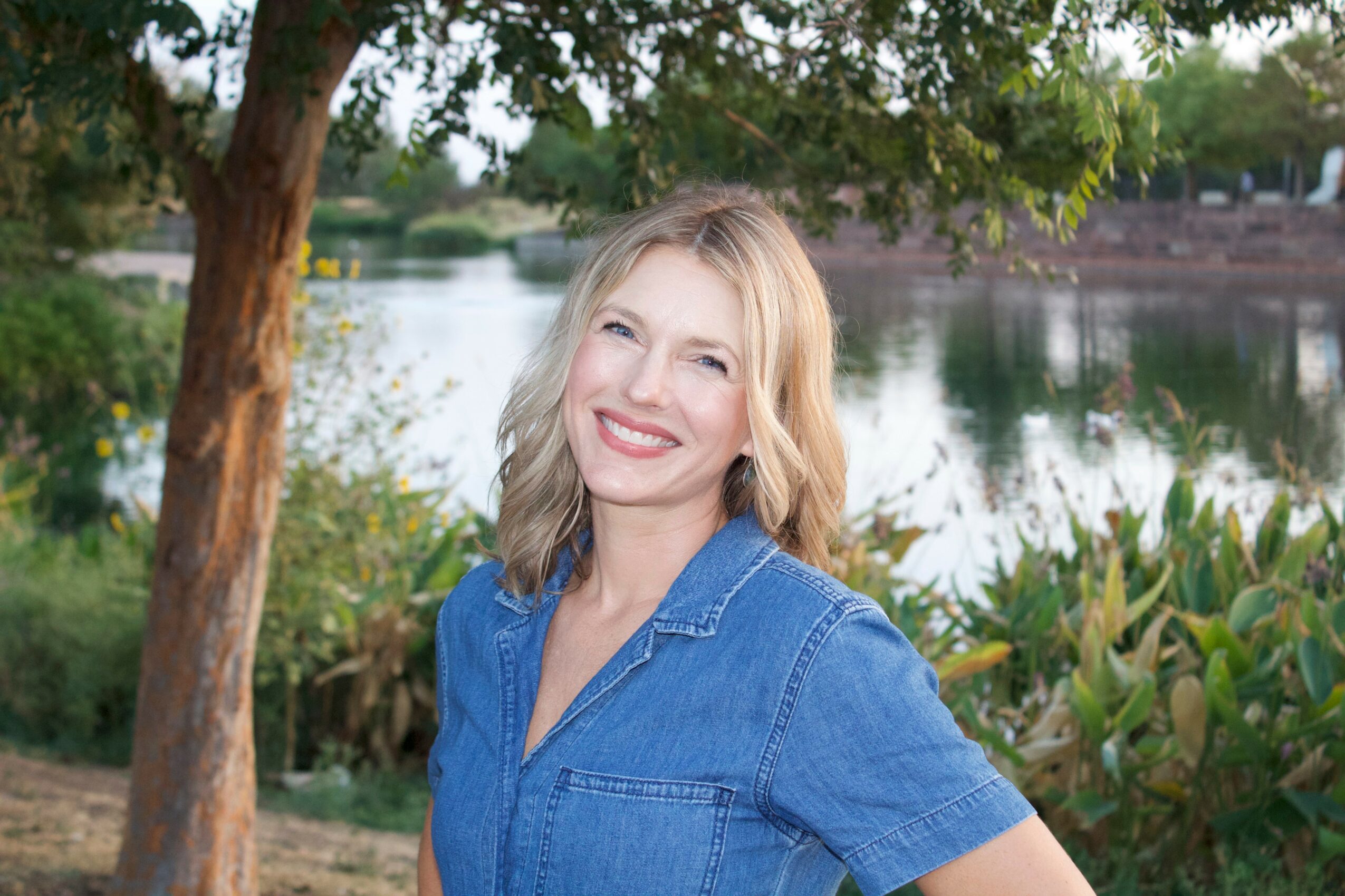 Smiling woman with blonde hair in a blue shirt outdoors near a body of water, trees, and greenery.