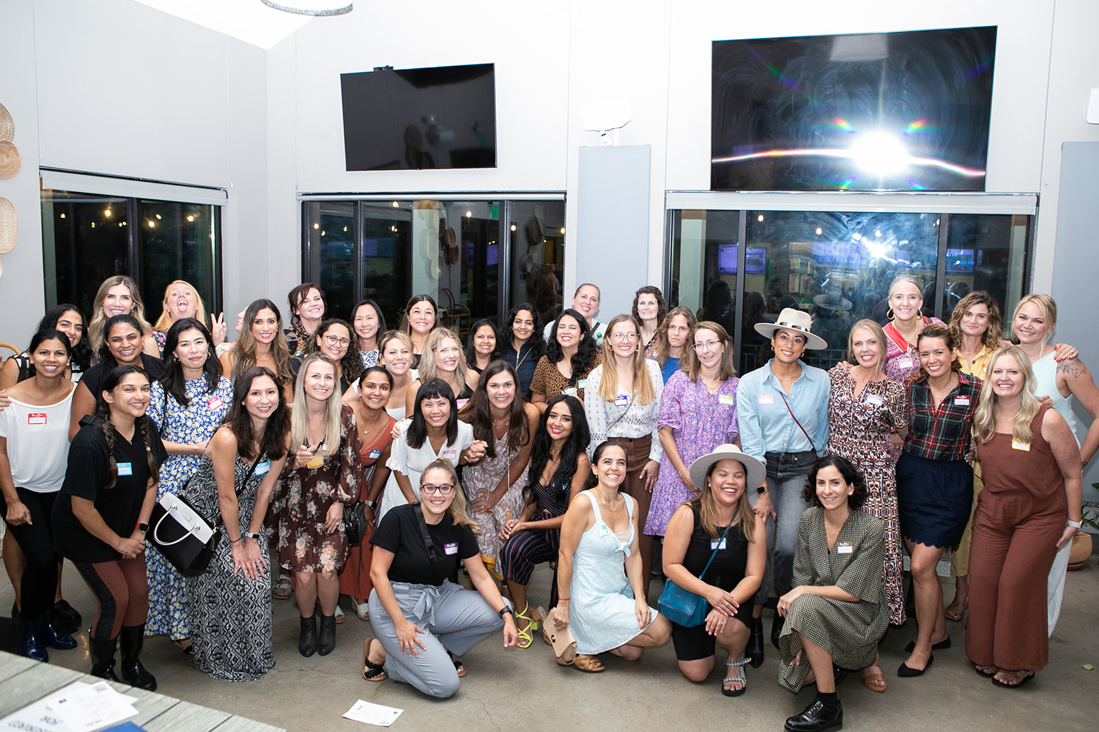 Group of women and girls gathered indoors, some smiling, in front of large windows and screens, with a space-themed image on the wall.