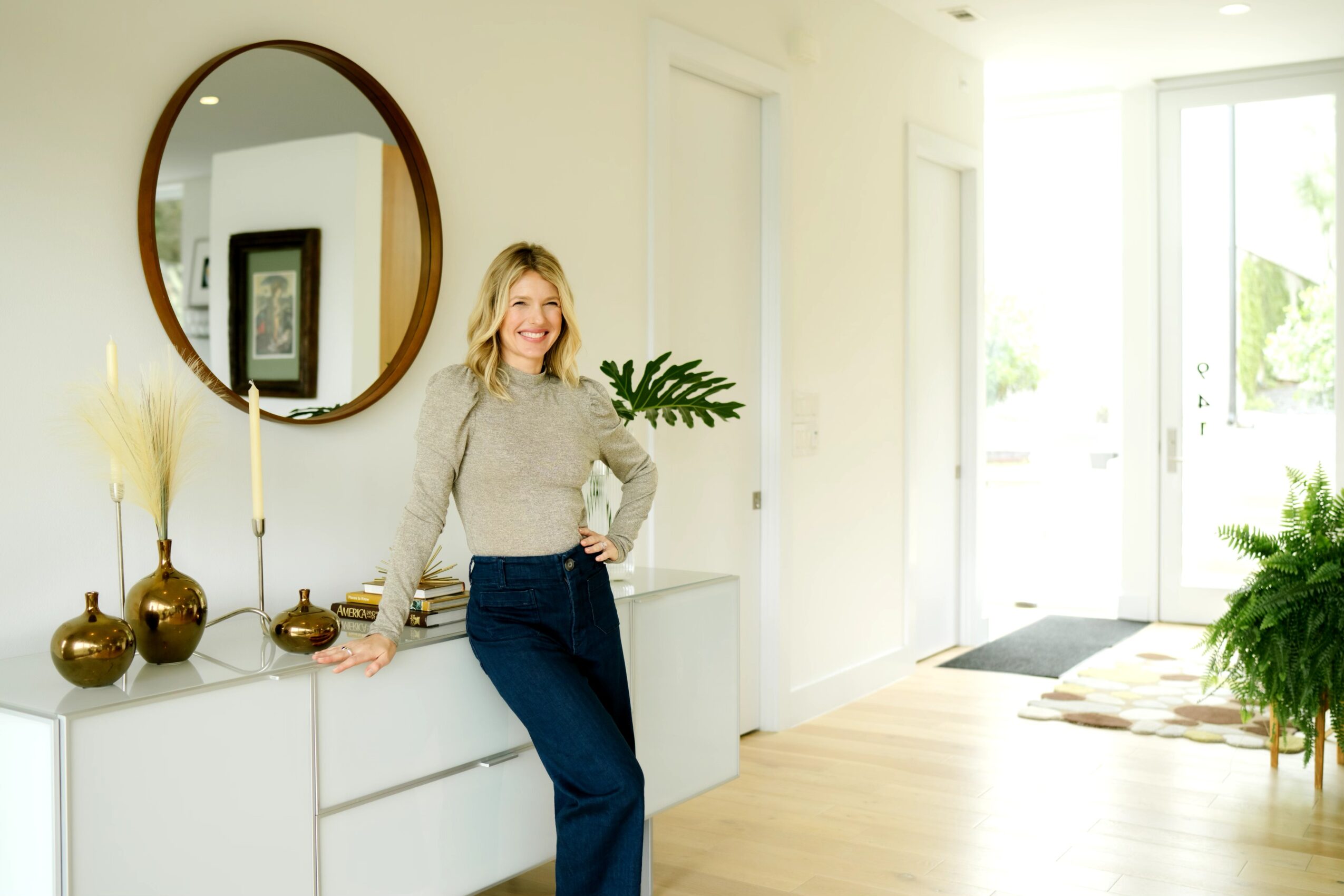 Woman with blonde hair smiling, standing in a bright, modern room with a mirror, plants, and decorative vases.