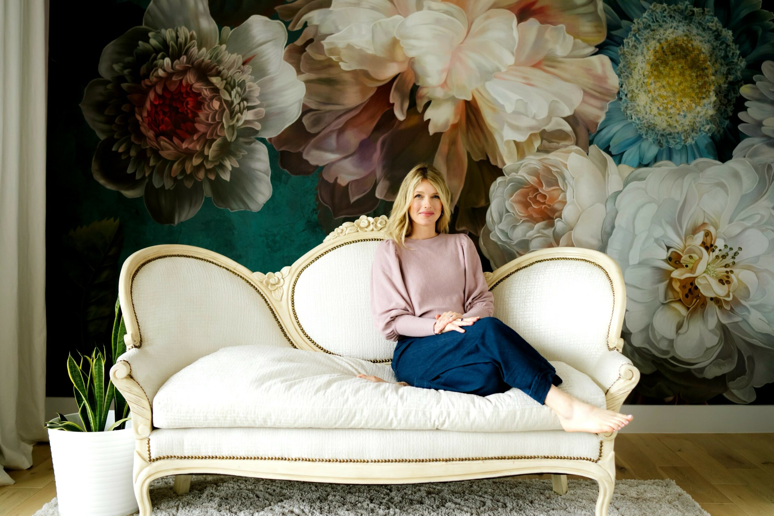 Woman sitting on a white vintage sofa with floral wallpaper background, wearing a pink top and blue skirt.