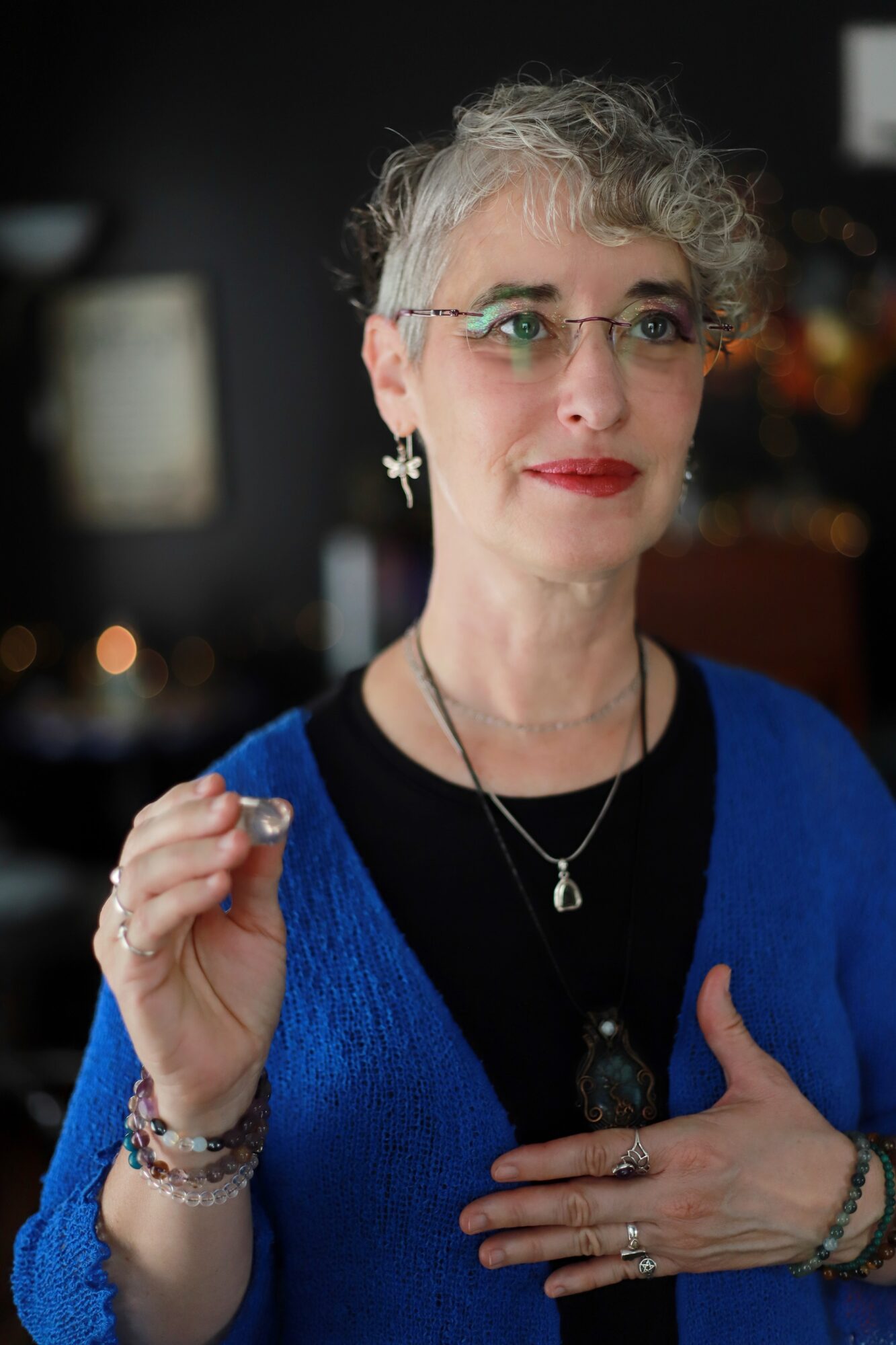 Woman with short curly gray hair, glasses, red lipstick, wearing a blue cardigan and jewelry, standing indoors.