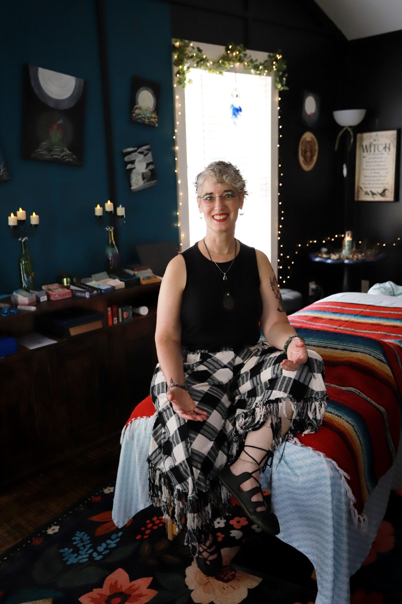 Woman with short curly hair and glasses sitting on a bed in a decorated room, smiling at the camera.