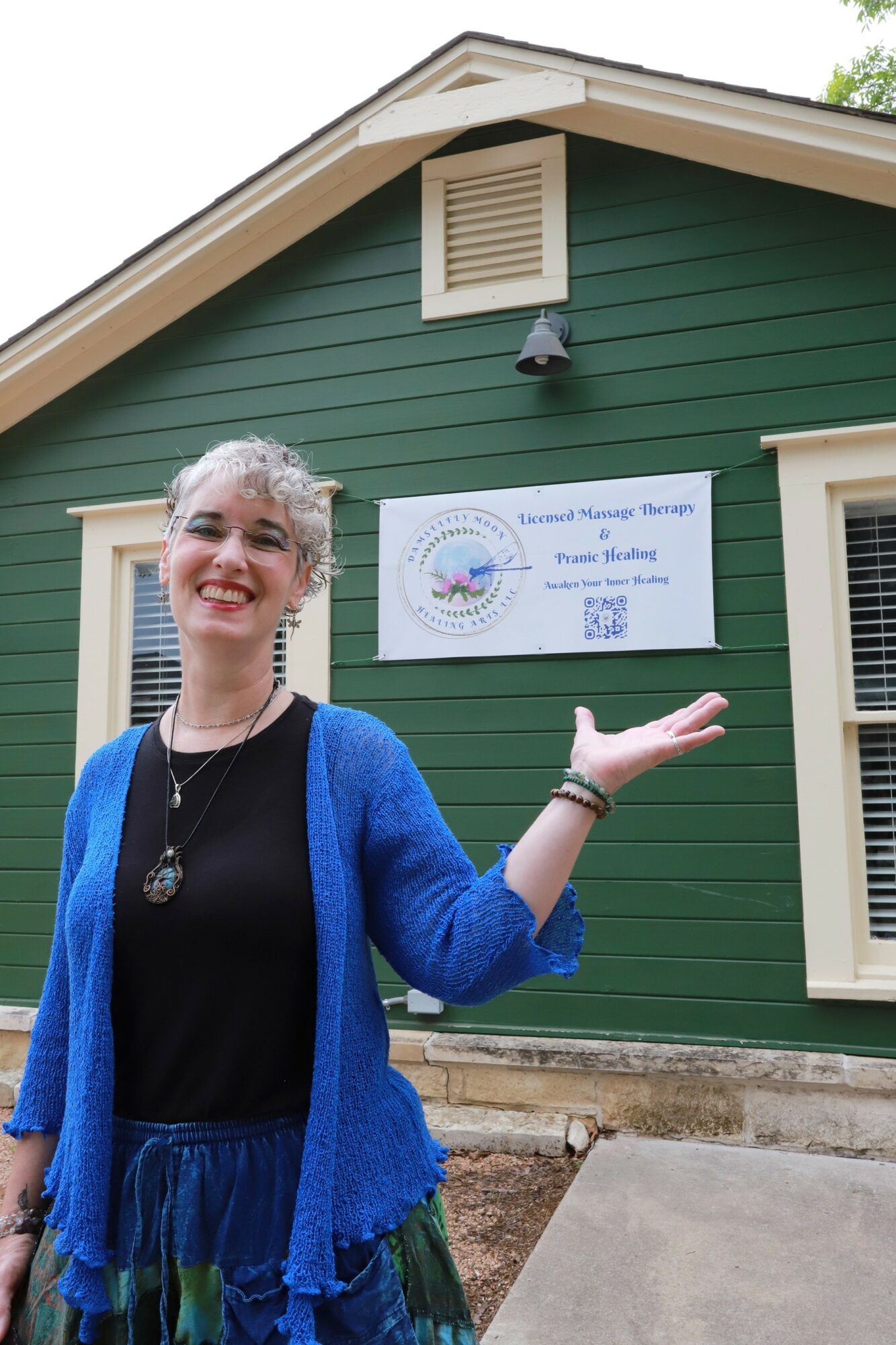 Smiling woman in blue cardigan gesturing outside a green building with a sign, overcast sky, concrete sidewalk, and windows.