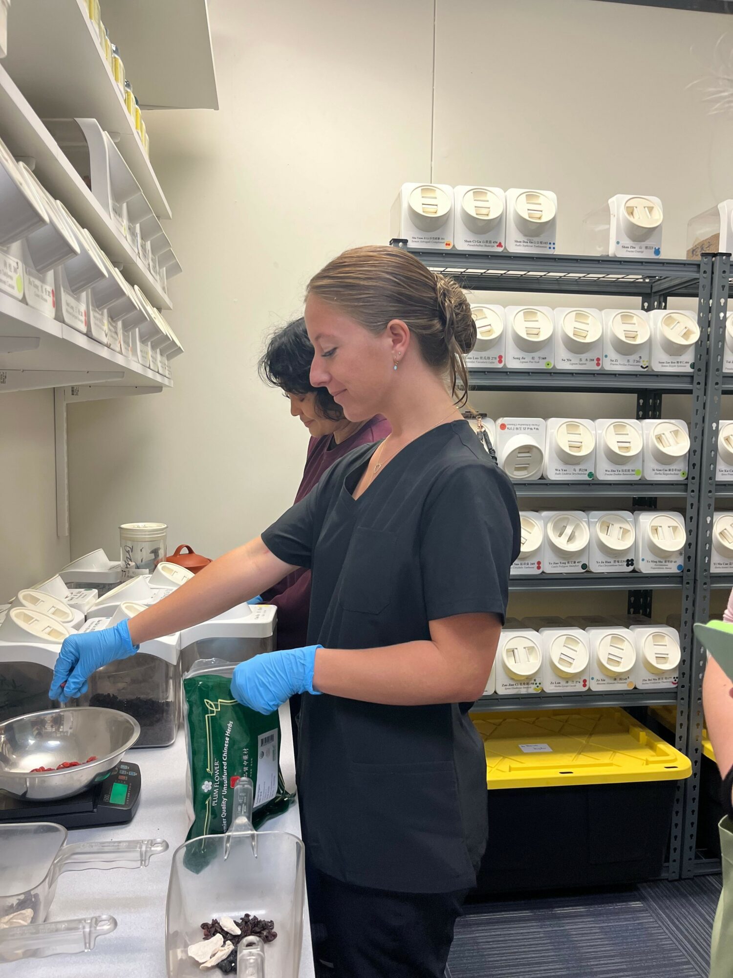 Woman in black scrubs and blue gloves working with medical supplies on a table, shelves with containers in background.